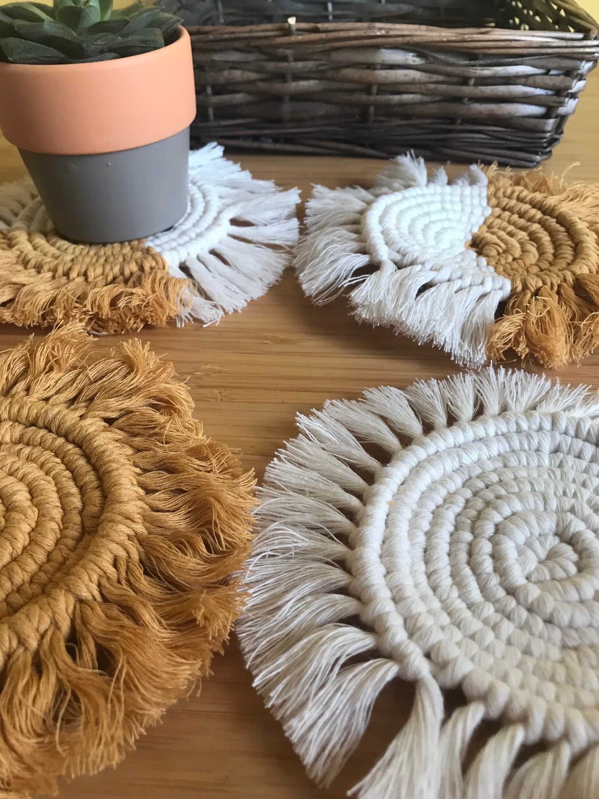 A close up image of a set of 4 round macrame coasters made with mustard yellow and natural yarns, with a soft fringed edge on a wooden table, with a small succulent houseplant and a brown woven basket.