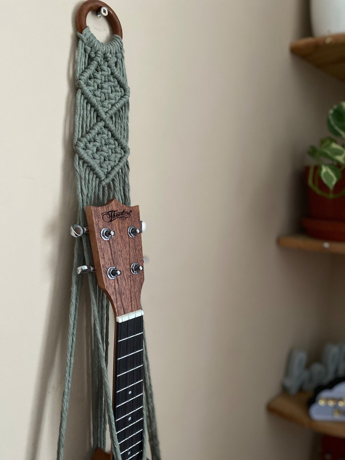 A dark brown ukulele sits inside of a handmade macrame wall hanging ukulele hanger, which is made with sage green recycled cotton, and a repurposed wooden hanging ring.