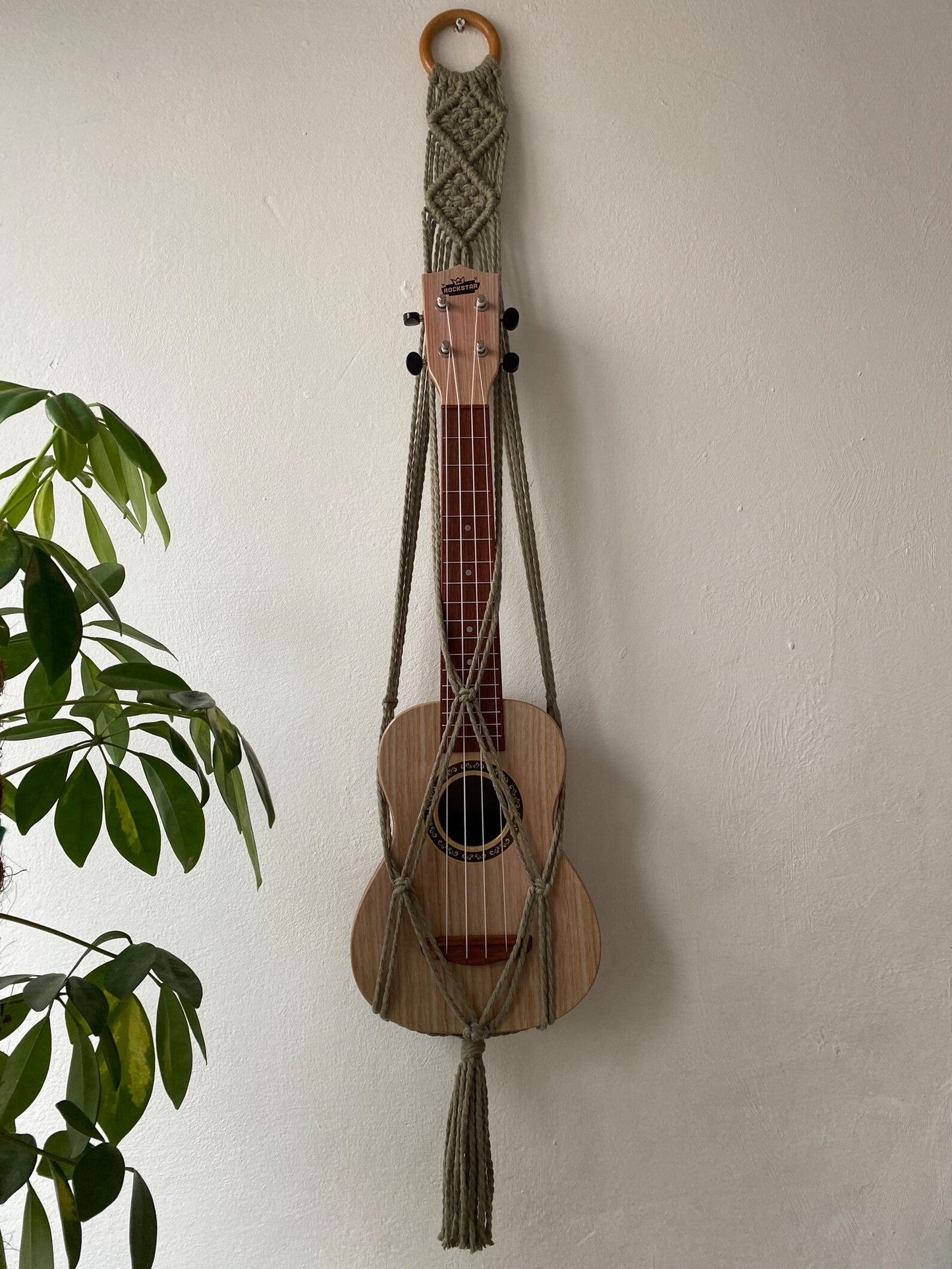 A light brown ukulele sits inside of a handmade macrame wall hanging ukulele hanger, which is made with sage green recycled cotton, and a repurposed wooden hanging ring. A large green leafy plant is to the left of the image.