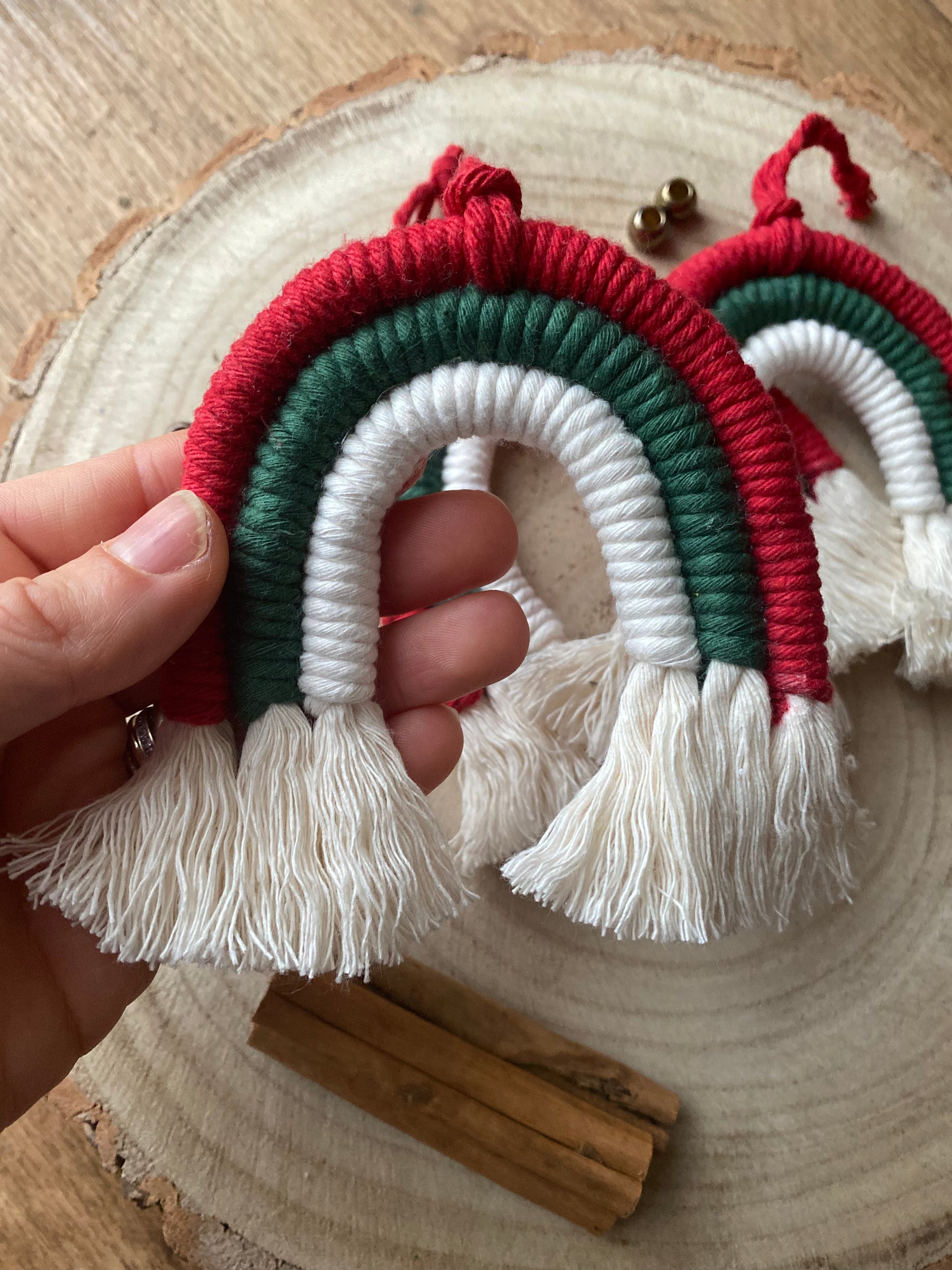 A hand holding one of a set of three handmade macrame rainbow Christmas decorations in red, green, and white yarn with white tassels, displayed on a wooden surface with cinnamon sticks for decoration.