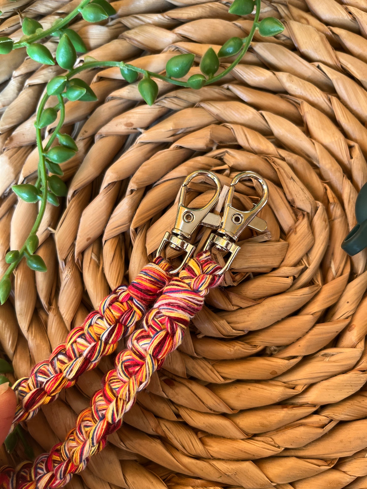 Handmade short macrame phone wristlet loop for phone case. Shown on a wicker mat. Handmade with multicoloured recycled cotton yarn, with a small metal lobster clasp on each end.