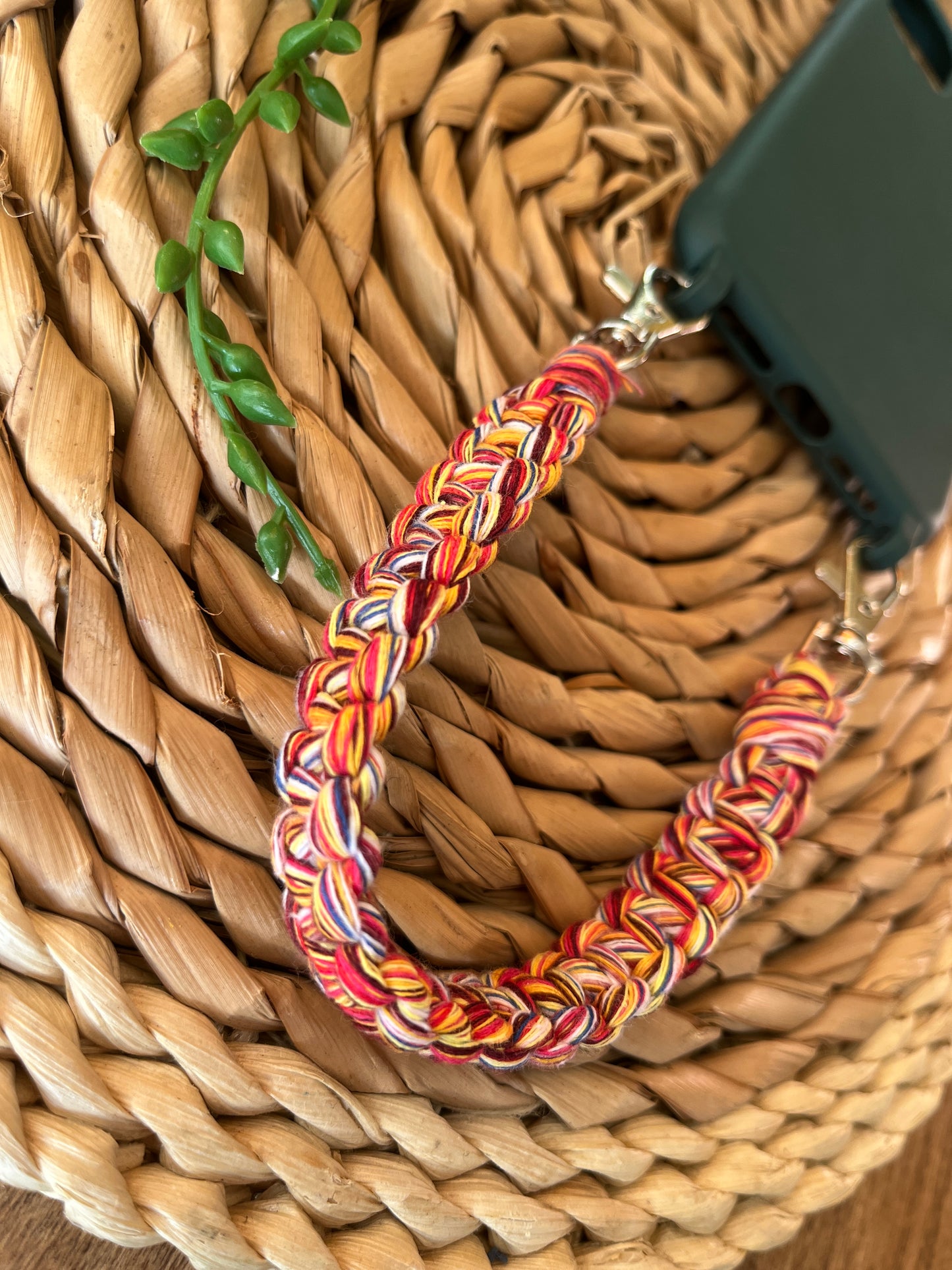 Handmade short macrame phone wristlet loop for phone case. Shown attached to a green phone case on a wicker mat. Handmade with multicoloured recycled cotton yarn, with a small metal lobster clasp on each end.
