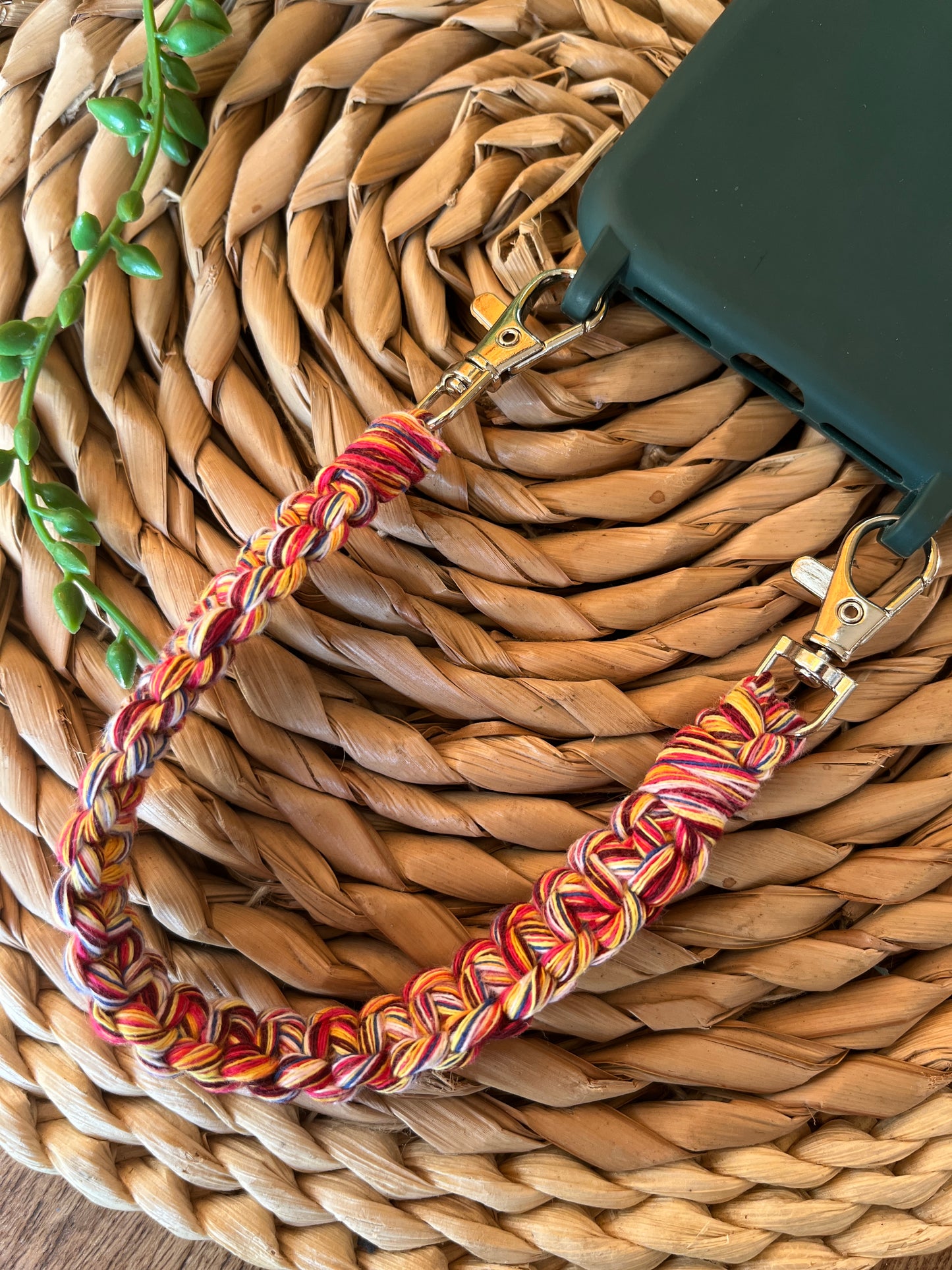 Handmade short macrame phone wristlet loop for phone case. Shown attached to a green phone case on a wicker mat. Handmade with multicoloured recycled cotton yarn, with a small metal lobster clasp on each end.