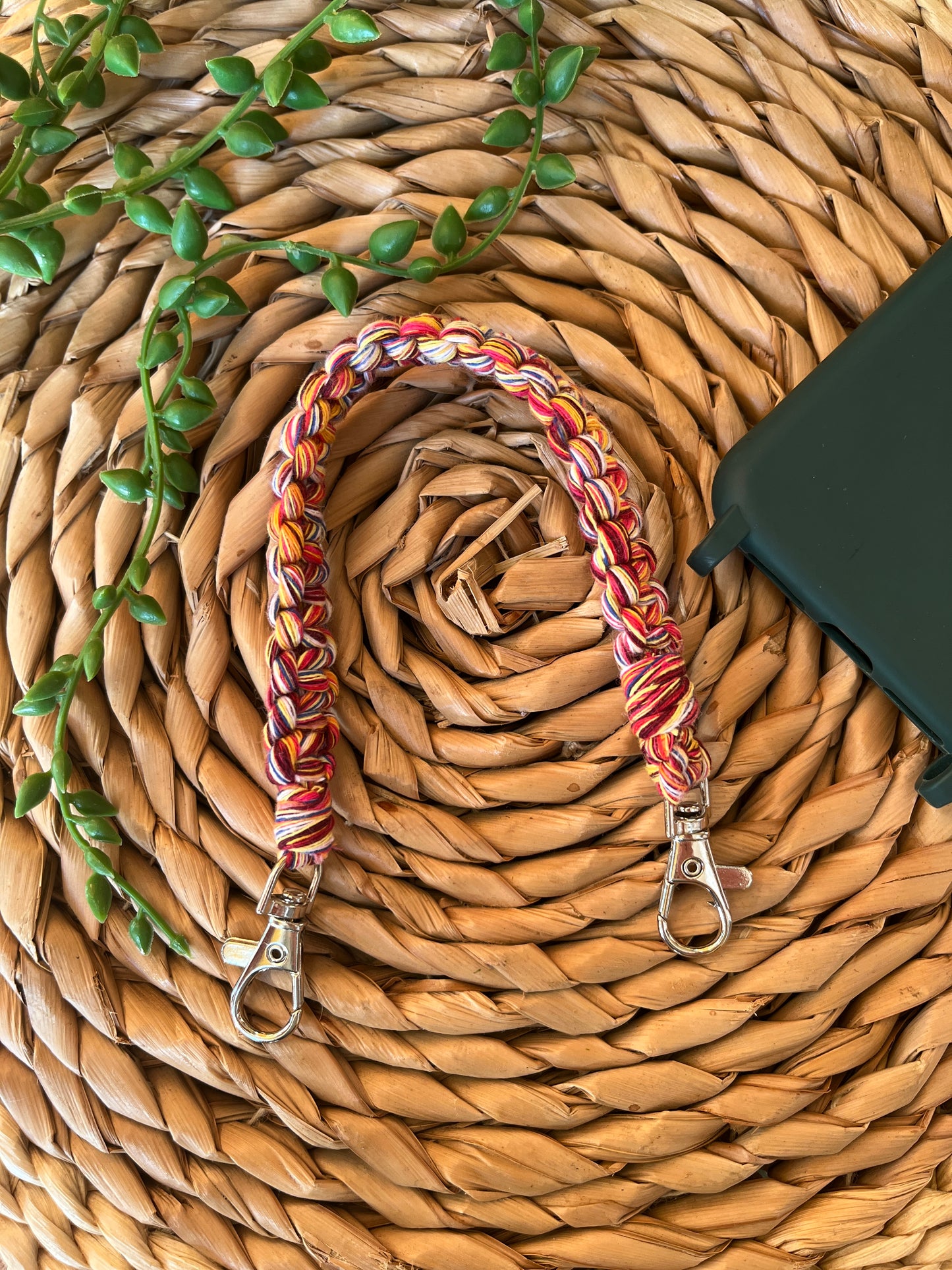 Handmade short macrame phone wristlet loop for phone case. Shown next to a green phone case on a wicker mat. Handmade with multicoloured recycled cotton yarn, with a small metal lobster clasp on each end.