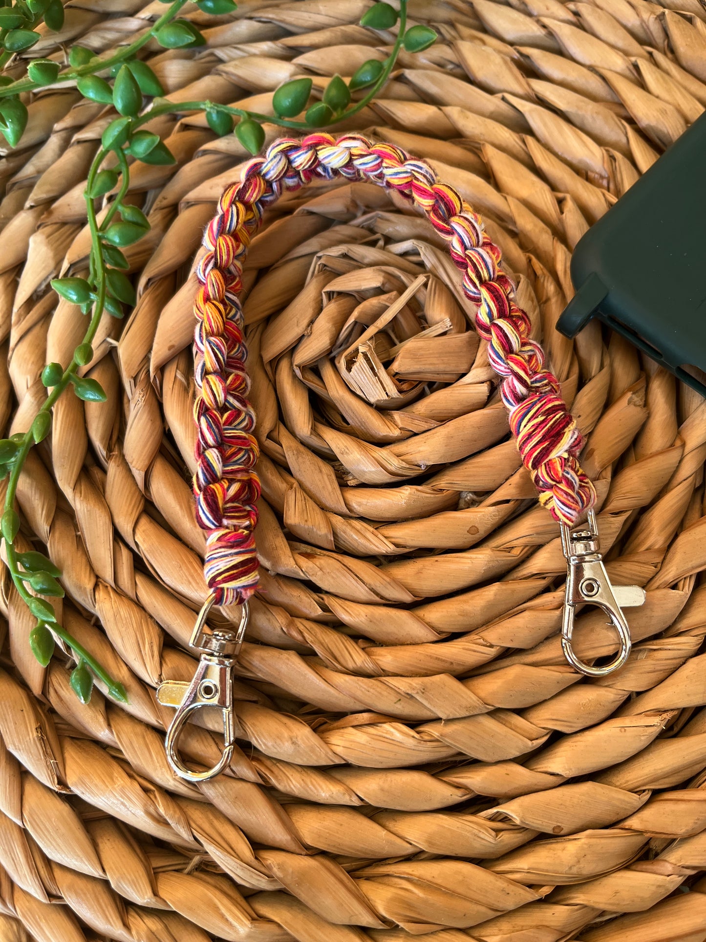 Handmade short macrame phone wristlet loop for phone case. Shown placed next to a green phone case on a wicker mat. Handmade with multicoloured recycled cotton yarn, with a small metal lobster clasp on each end.
