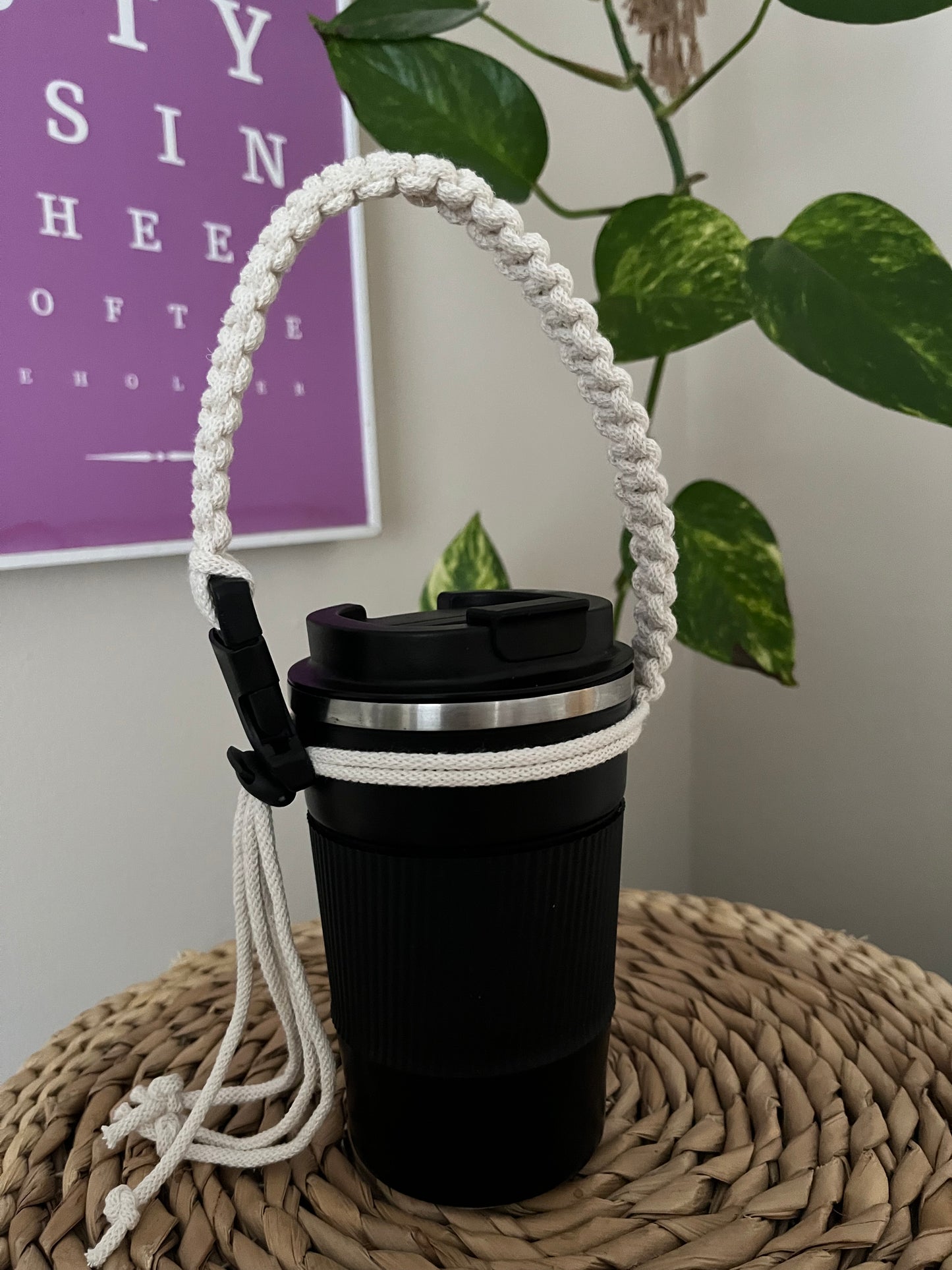 A handmade macrame knotted coffee cup holder strap, made with natural eco friendly recycled cotton, with a black buckle and toggle fastening is attached to a black travel cup, with a long green plant in the background.