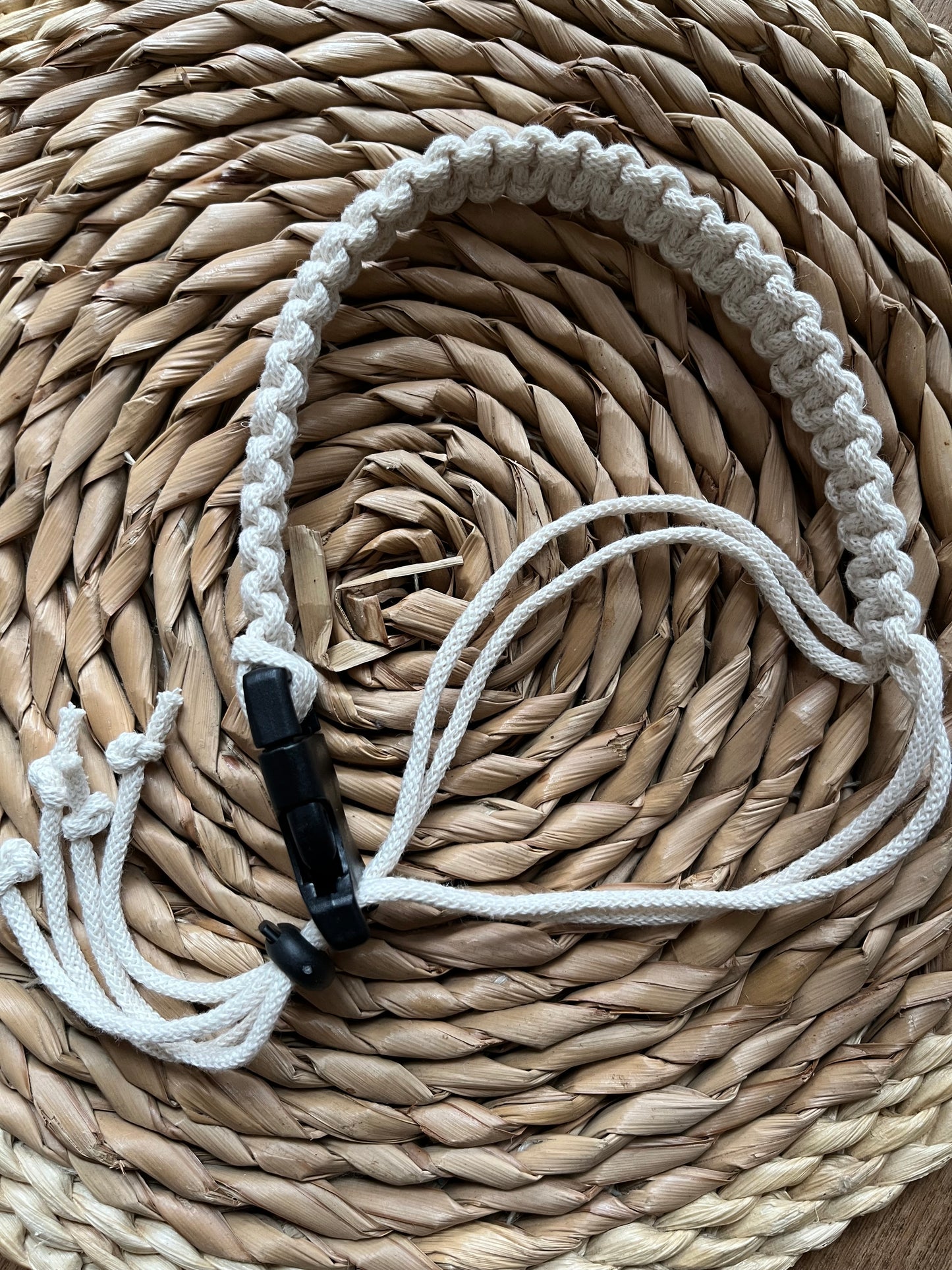 A handmade macrame adjustable coffee cup holder strap is laid out on a wicker surface. The strap features neat knots, and a black buckle and toggle fastening.