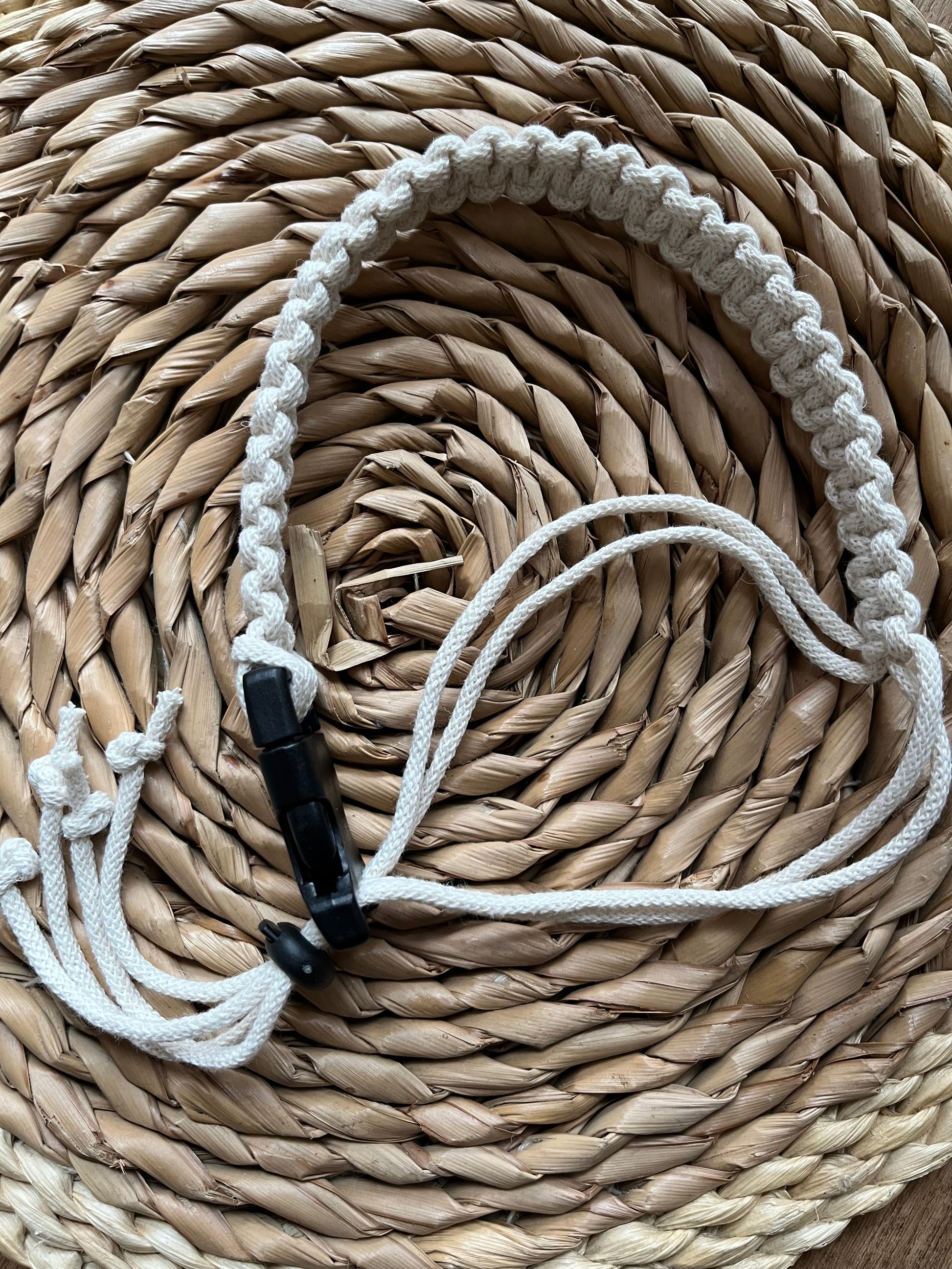 A handmade macrame adjustable coffee cup holder strap is laid out on a wicker surface. The strap features neat knots, and a black buckle and toggle fastening.