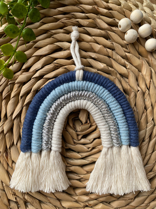 Handmade blue macramé rainbow wall hanging made with recycled cotton cord in soft blue and neutral tones, shown against a wicker background.