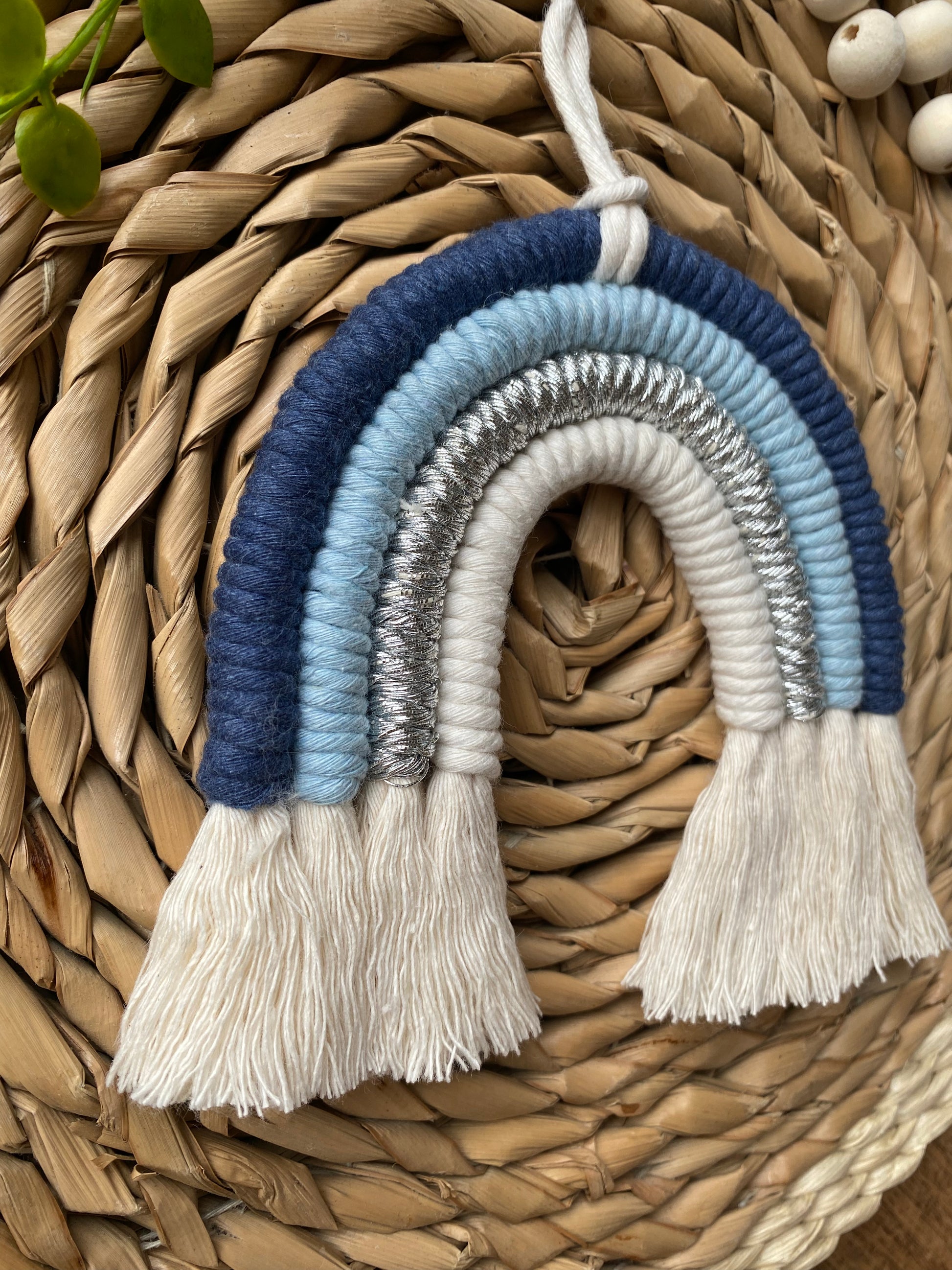 Close up of a handmade blue macramé rainbow wall hanging made with recycled cotton cord in soft blue and neutral tones, shown against a wicker background.