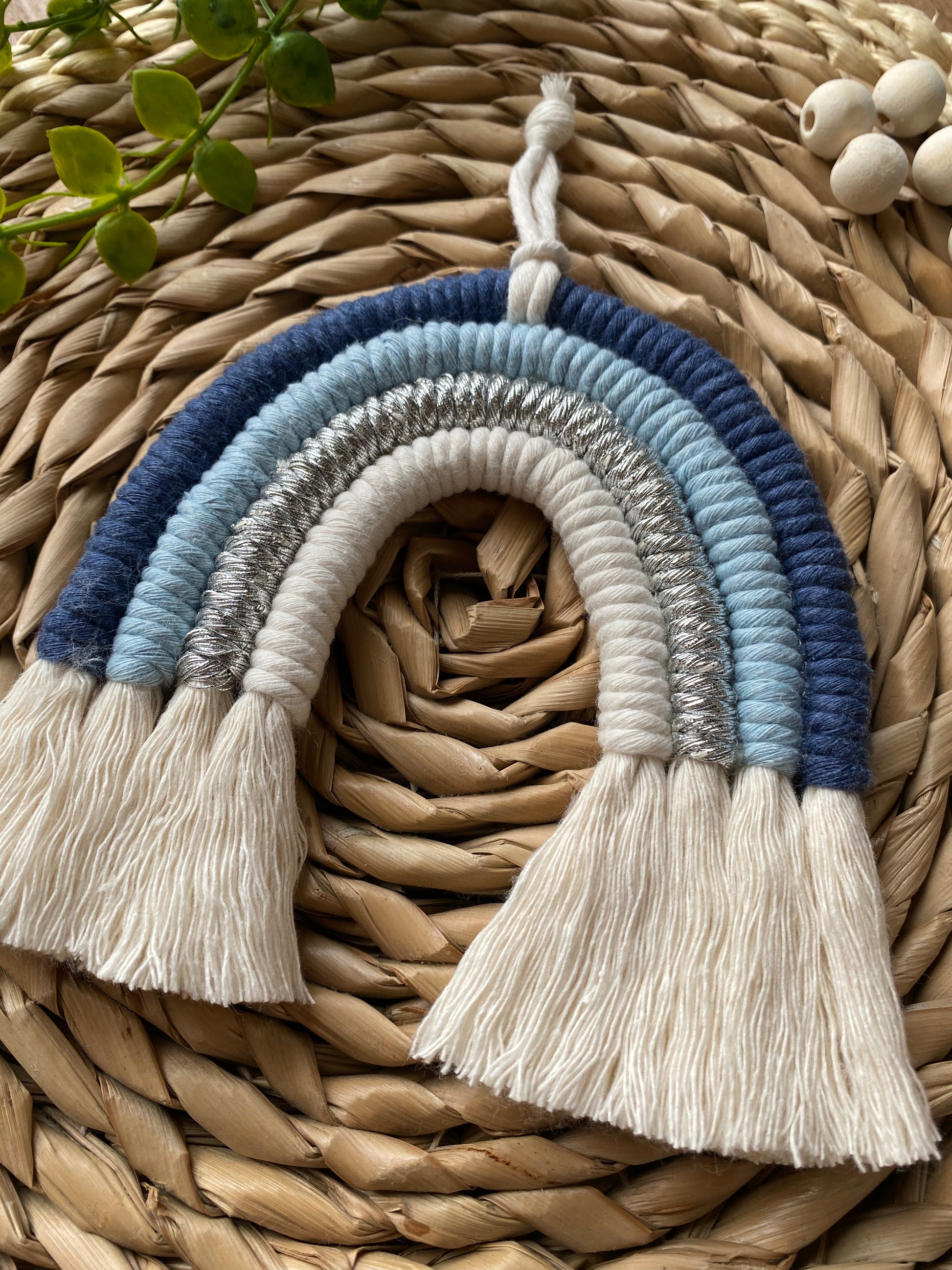 A handmade small blue macrame rainbow  from Macra-Made-With-Love made from blue, silver, and white yarn is displayed on a woven rattan surface.