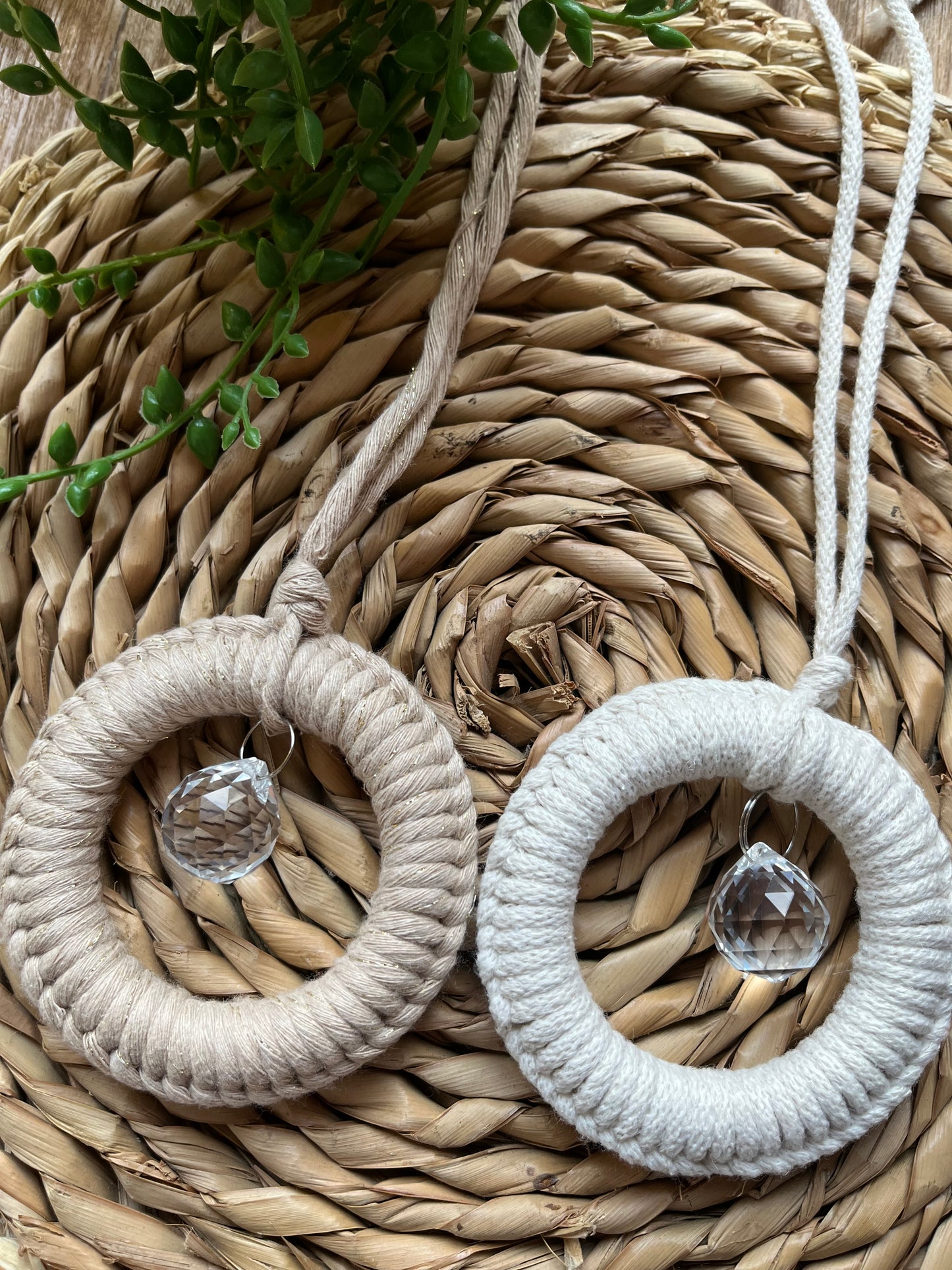 Two handmade macrame circular hanging suncatchers, made with recycled cotton with a small crystal prism. The suncatchers are on a wicker surface with some green plant vines.