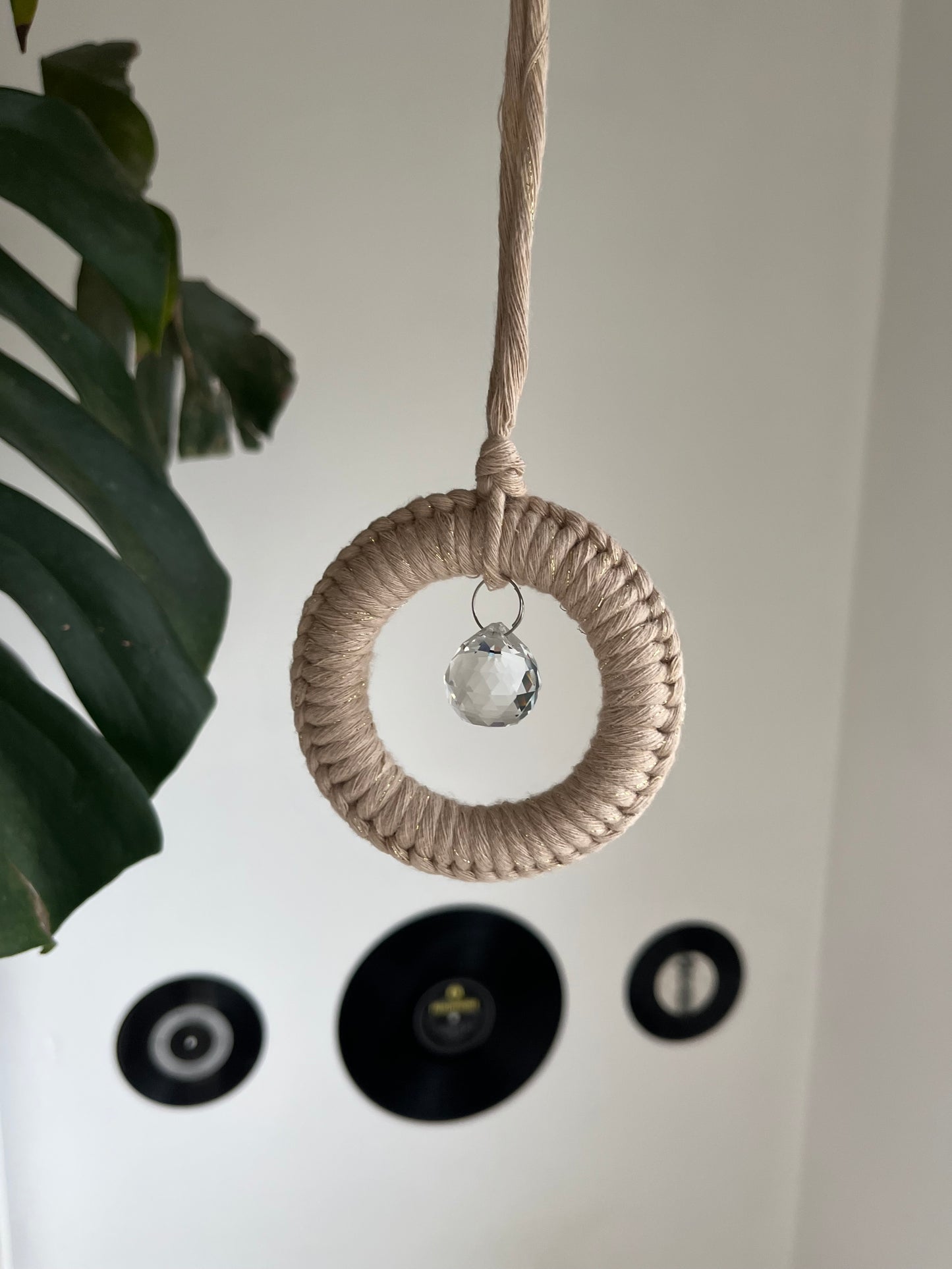 A handmade macrame circular hanging suncatcher, made with recycled cotton in sparkly gold with a small crystal prism. There are records hanging on a white wall, and a green leaf in the background.