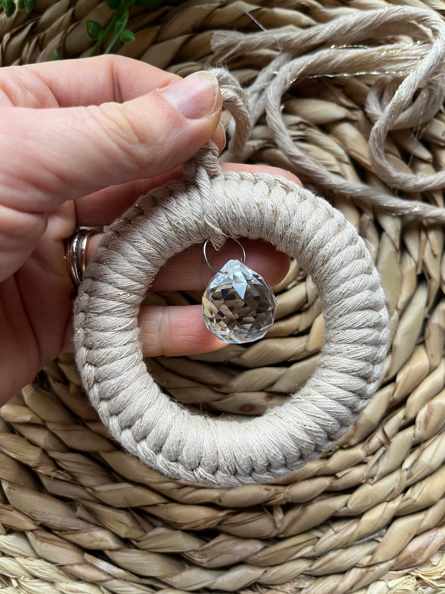 A handmade macrame circular hanging suncatcher, made with recycled cotton in a sparkly gold colour, with a small crystal prism. The suncatcher is on a wicker surface.
