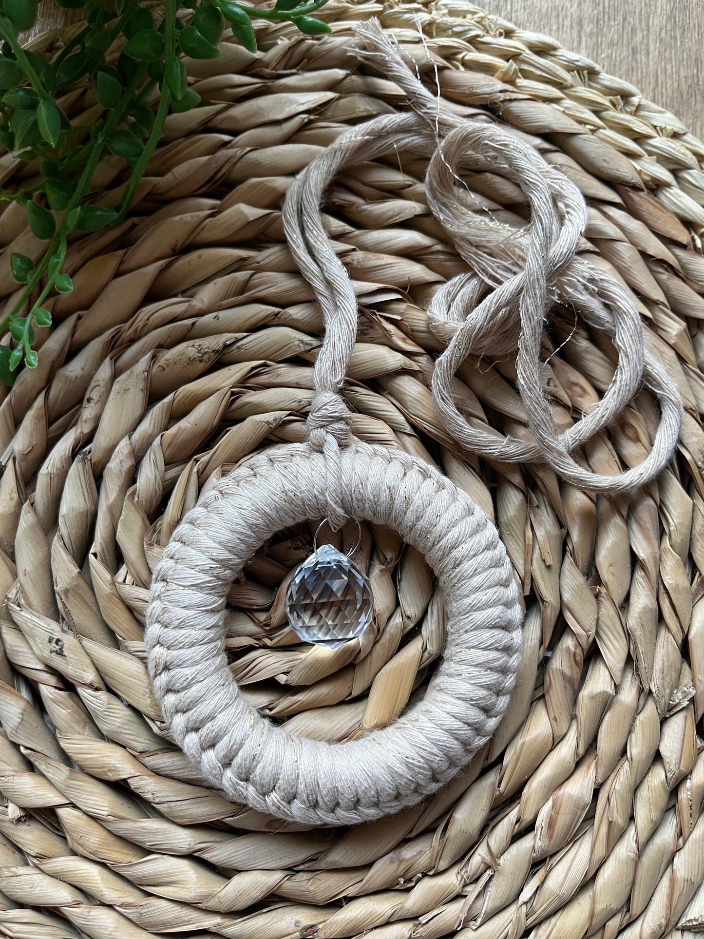 A handmade macrame circular hanging suncatcher, made with recycled cotton in sparkly gold, with a small crystal prism. The suncatcher is on a wicker surface with some green plant vines.