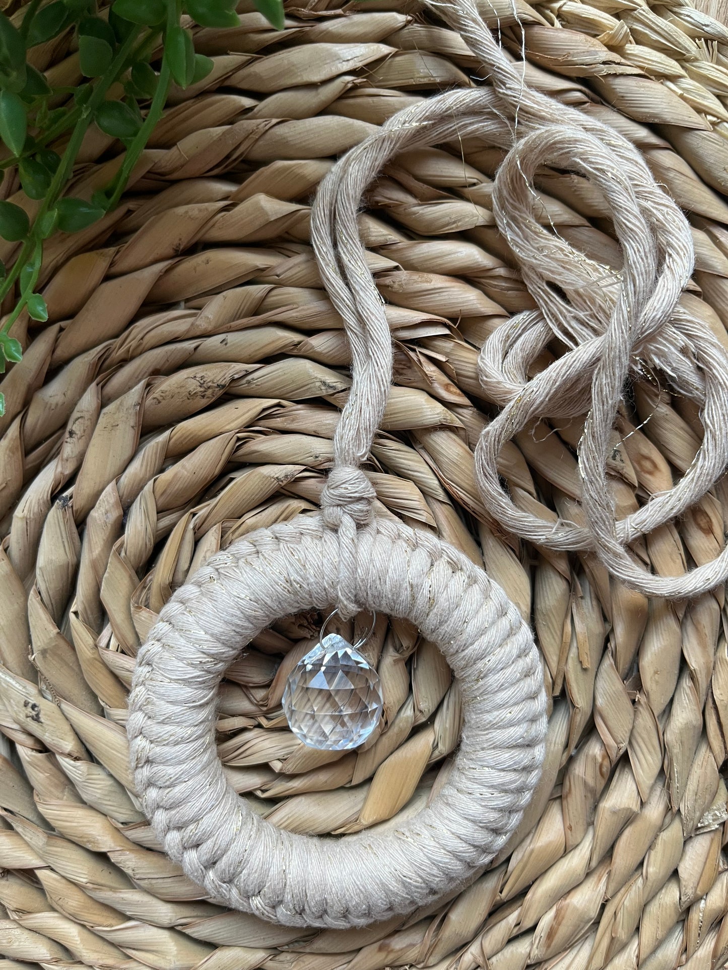 A handmade macrame circular hanging suncatcher, made with recycled cotton in sparkly gold, with a small crystal prism. The suncatcher is on a wicker surface with some green plant vines.