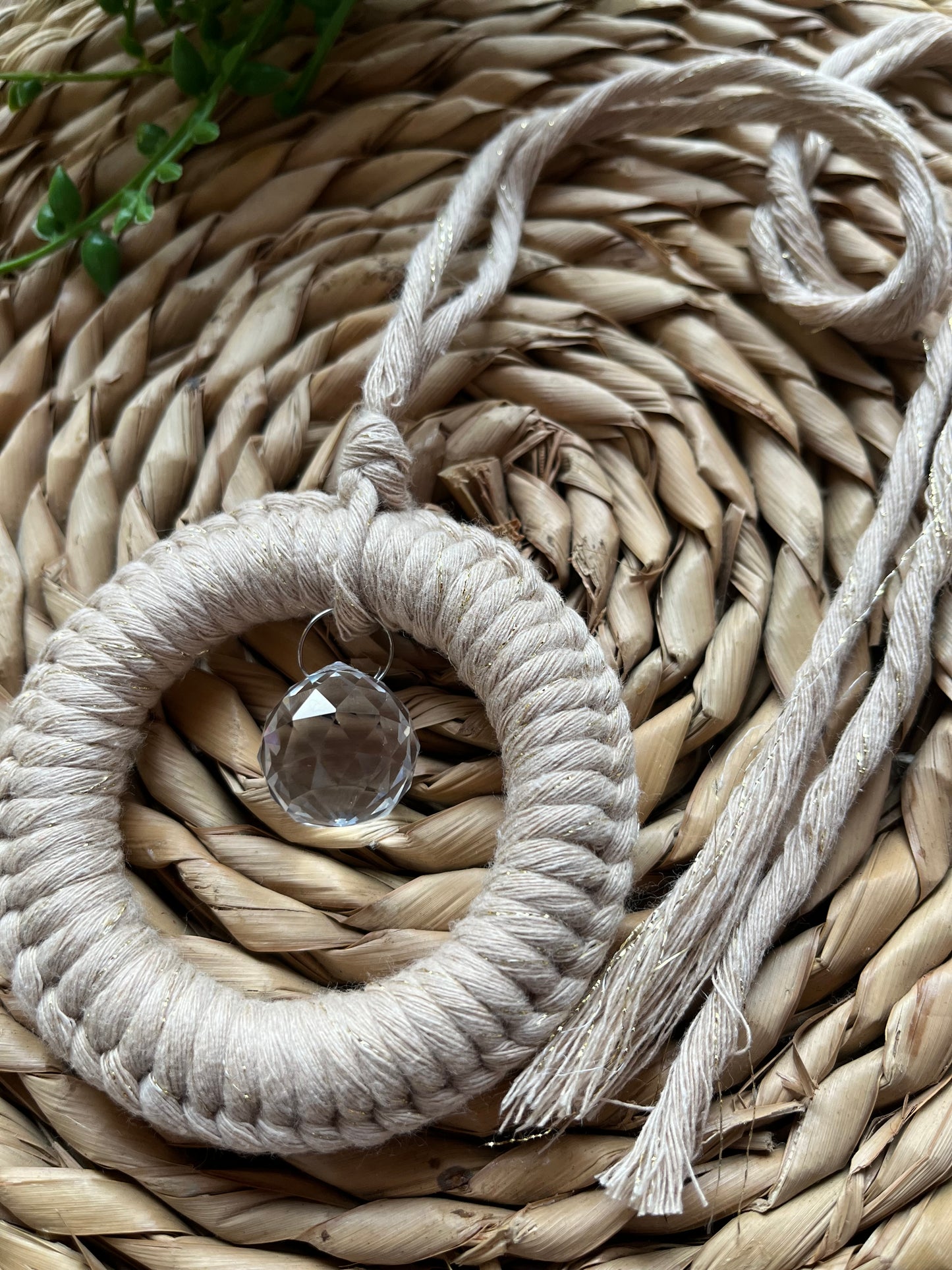 A handmade macrame circular hanging suncatcher, made with recycled cotton in sparkly gold, with a small crystal prism. The suncatcher is on a wicker surface with some green plant vines.