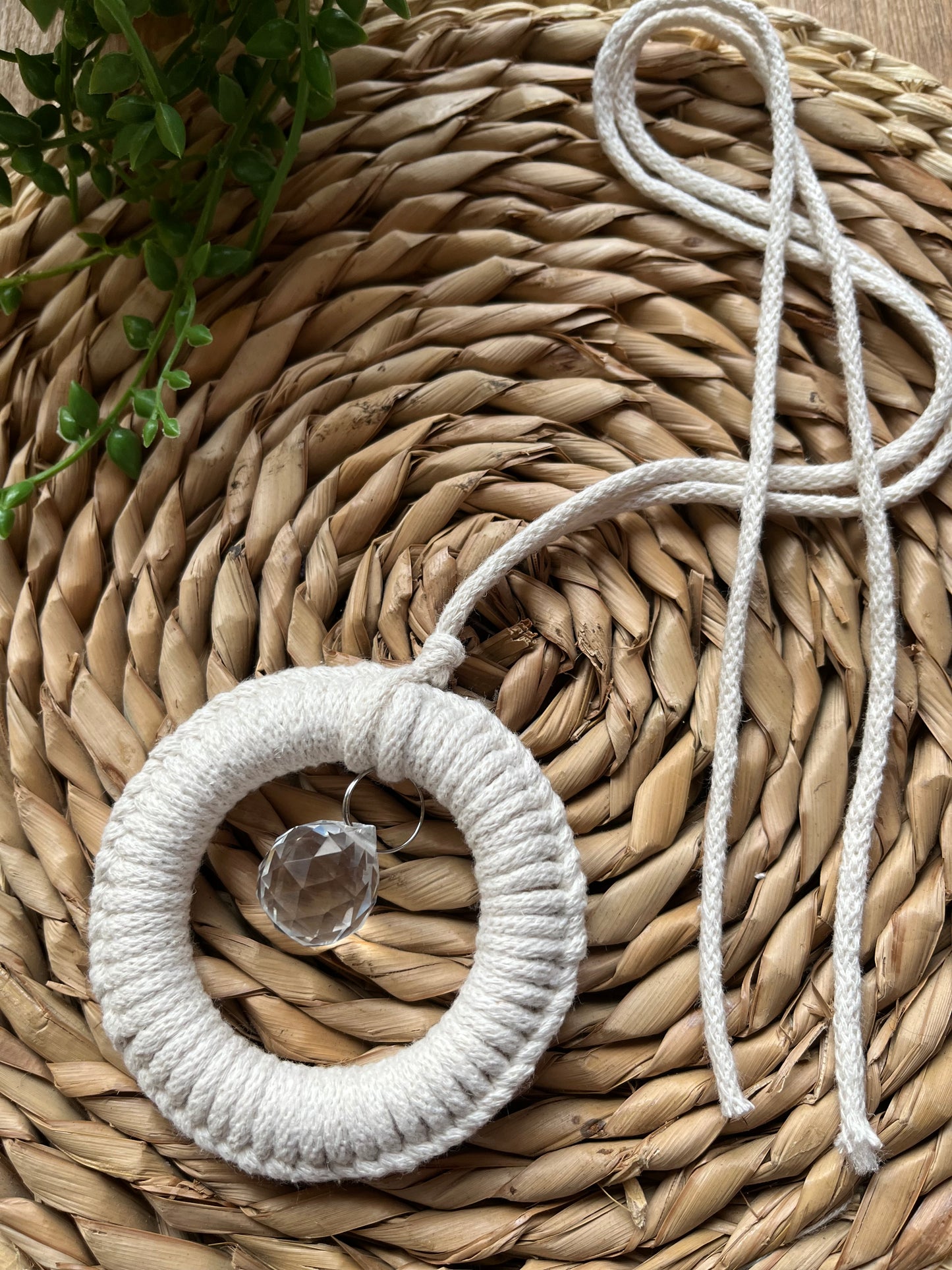 A handmade macrame circular hanging suncatcher, made with recycled cotton in an off white natural colour, with a small crystal prism. The suncatcher is on a wicker surface with some green plant vines.