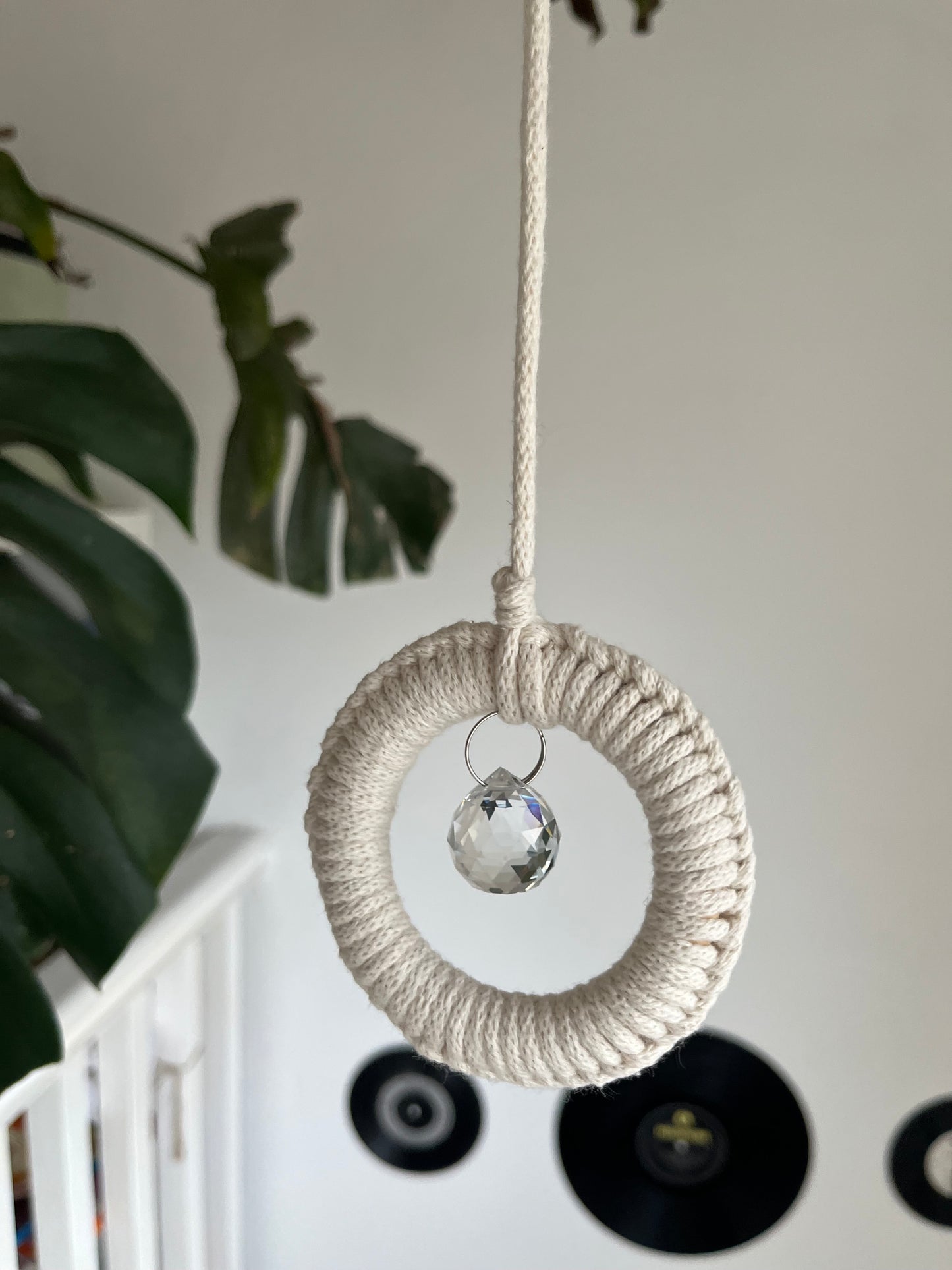 A handmade macrame circular hanging suncatcher, made with recycled cotton in an of white colour, with a small crystal prism. There is a green leafy plant in the background.