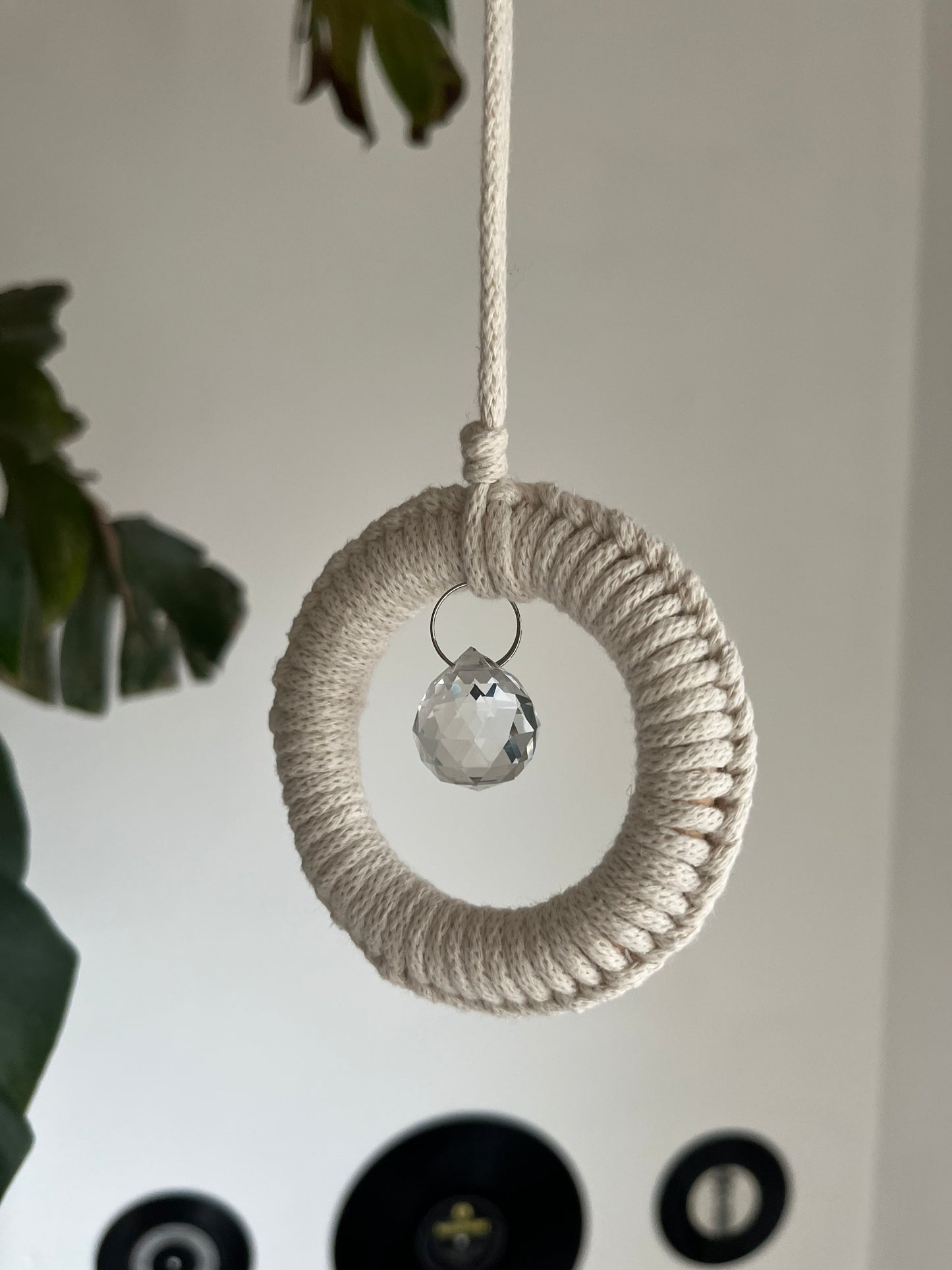 A handmade macrame circular hanging suncatcher, made with recycled cotton in an of white colour, with a small crystal prism. There is a green leafy plant in the background.