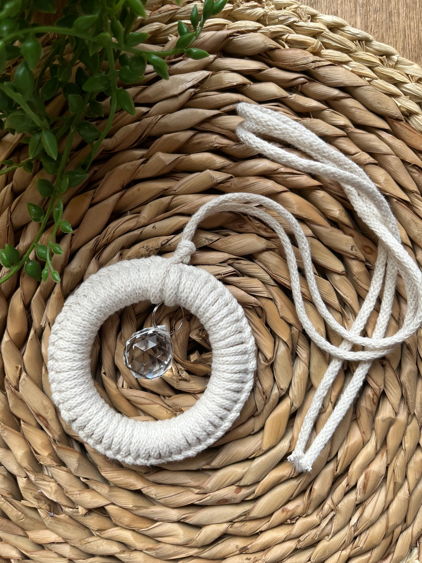 A handmade macrame circular hanging suncatcher, made with recycled cotton in an off white natural colour with a small crystal prism. The suncatcher is laid on a round wicker surface.