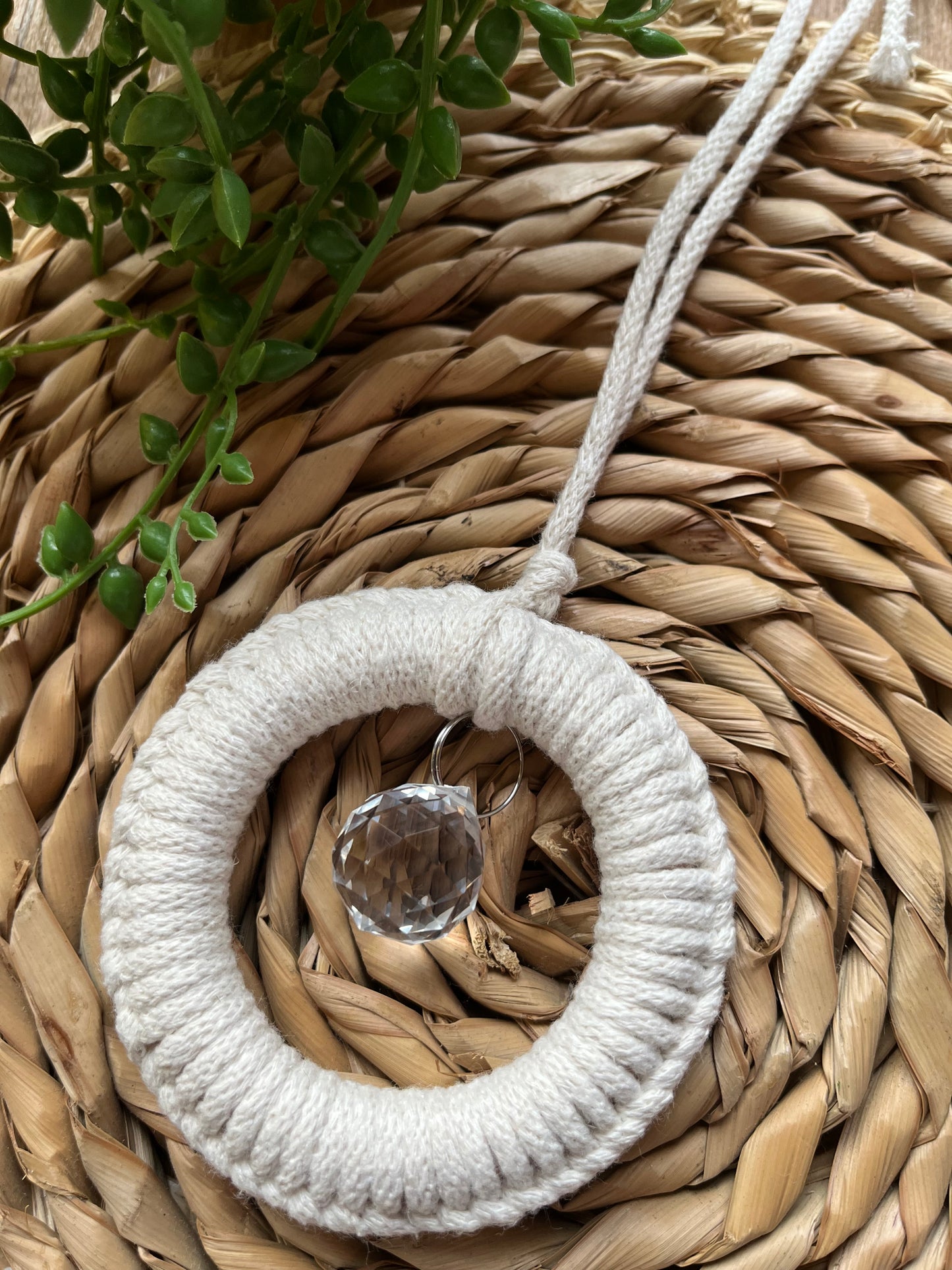 A handmade macrame circular hanging suncatcher, made with recycled cotton in an off white natural colour, with a small crystal prism. The suncatcher is on a wicker surface with some green plant vines.