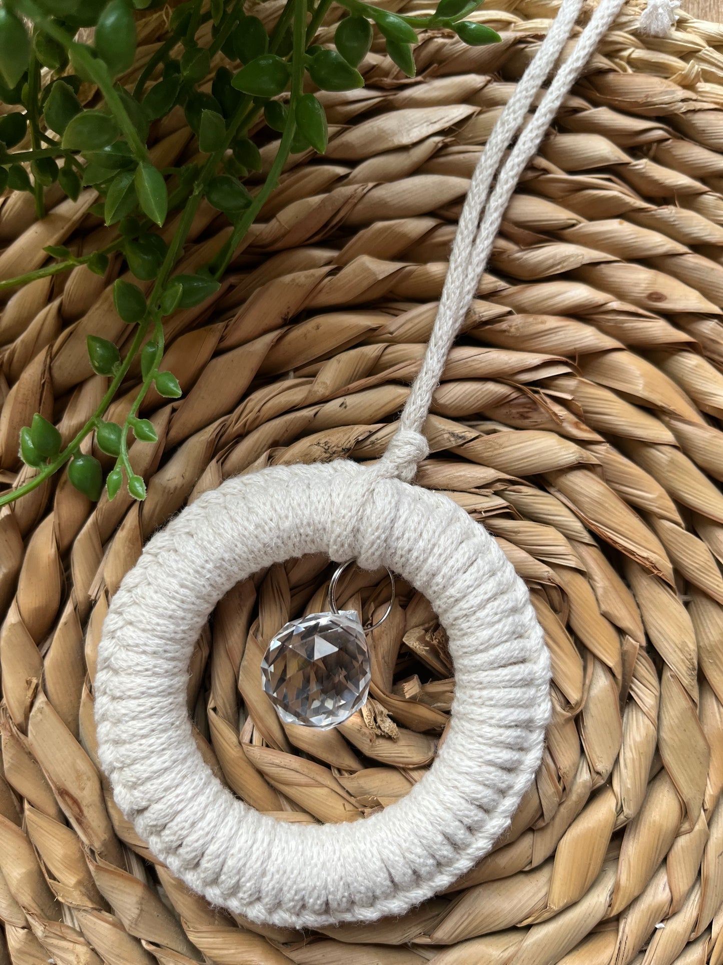 A handmade macrame circular hanging suncatcher, made with recycled cotton in an off white natural colour, with a small crystal prism. The suncatcher is on a wicker surface with some green plant vines.