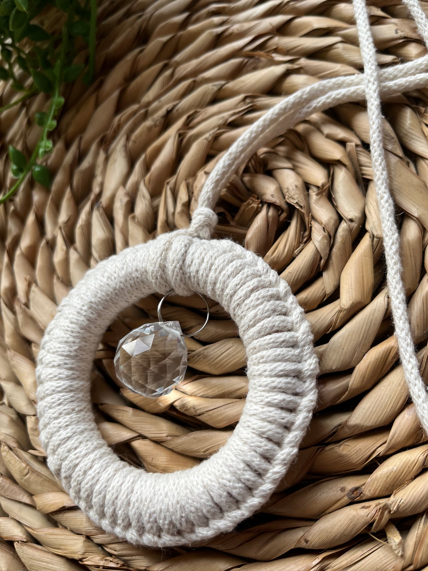 A handmade macrame circular hanging suncatcher, made with recycled cotton in an off white natural colour, with a small crystal prism. The suncatcher is on a wicker surface with some green plant vines.