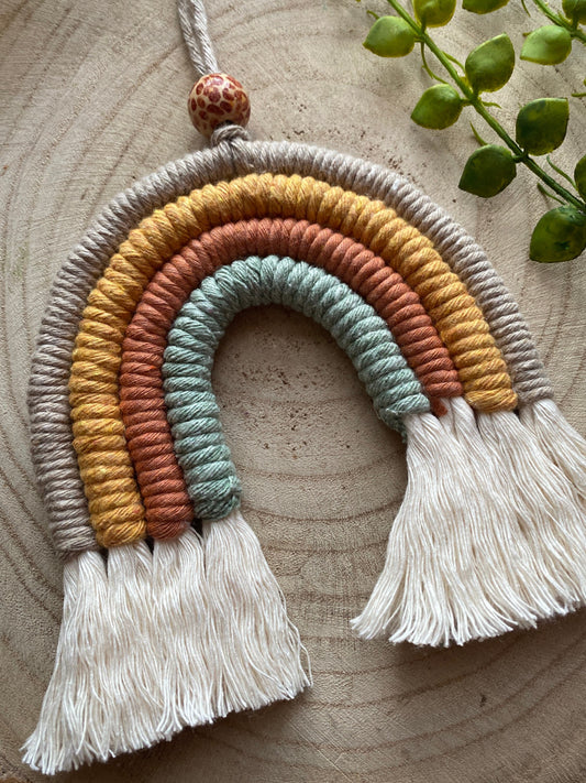 A handmade macrame rainbow, made with neutral shades of beige, yellow, rust and green yarns, with fluffy white tassels, and a patterned wooden bead, laid on a wooden surface with some green plant vines.