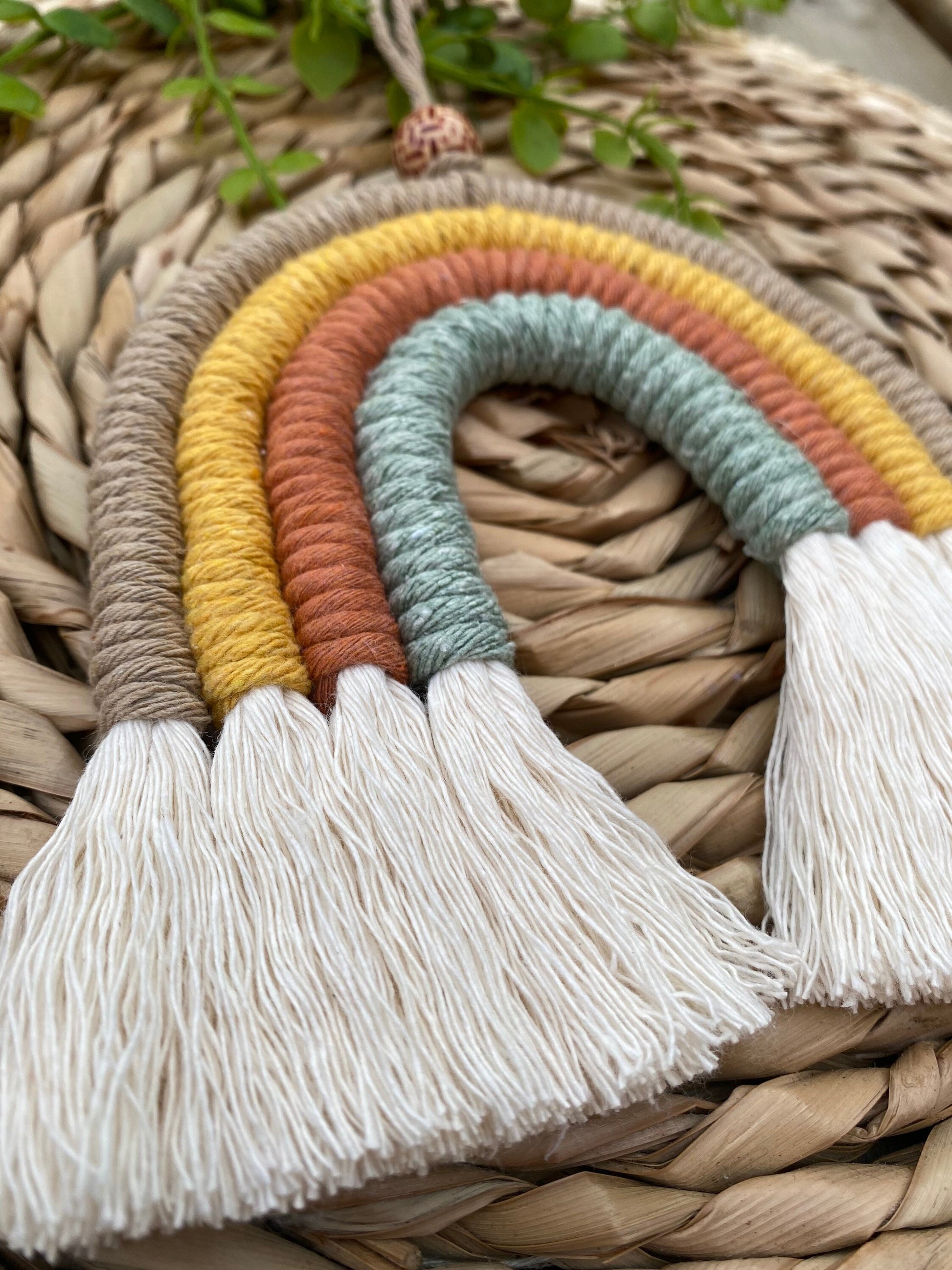 A side angled close up view of a handmade macrame rainbow, made with neutral shades of beige, yellow, rust and green yarns, with fluffy white tassels, and a patterned wooden bead, laid on a wicker mat.