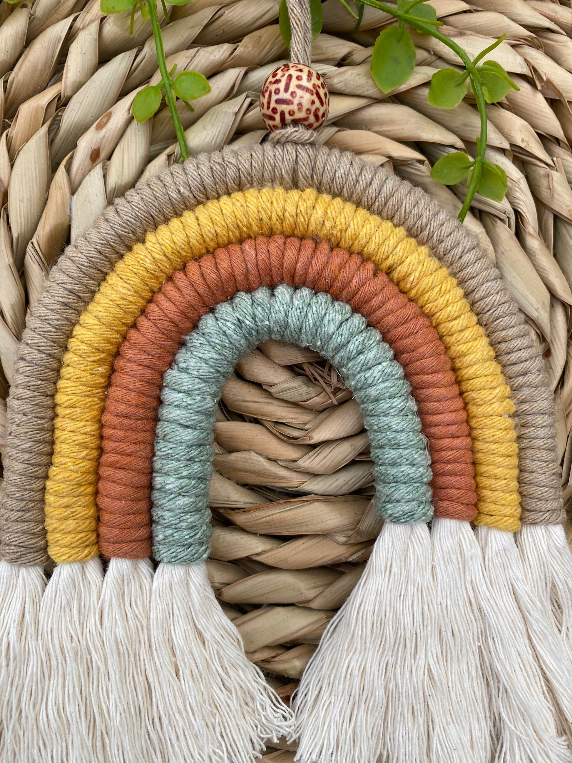 A close up of a handmade macrame rainbow, made with neutral shades of beige, yellow, rust and green yarns, with fluffy white tassels, and a patterned wooden bead, laid on a wicker mat.