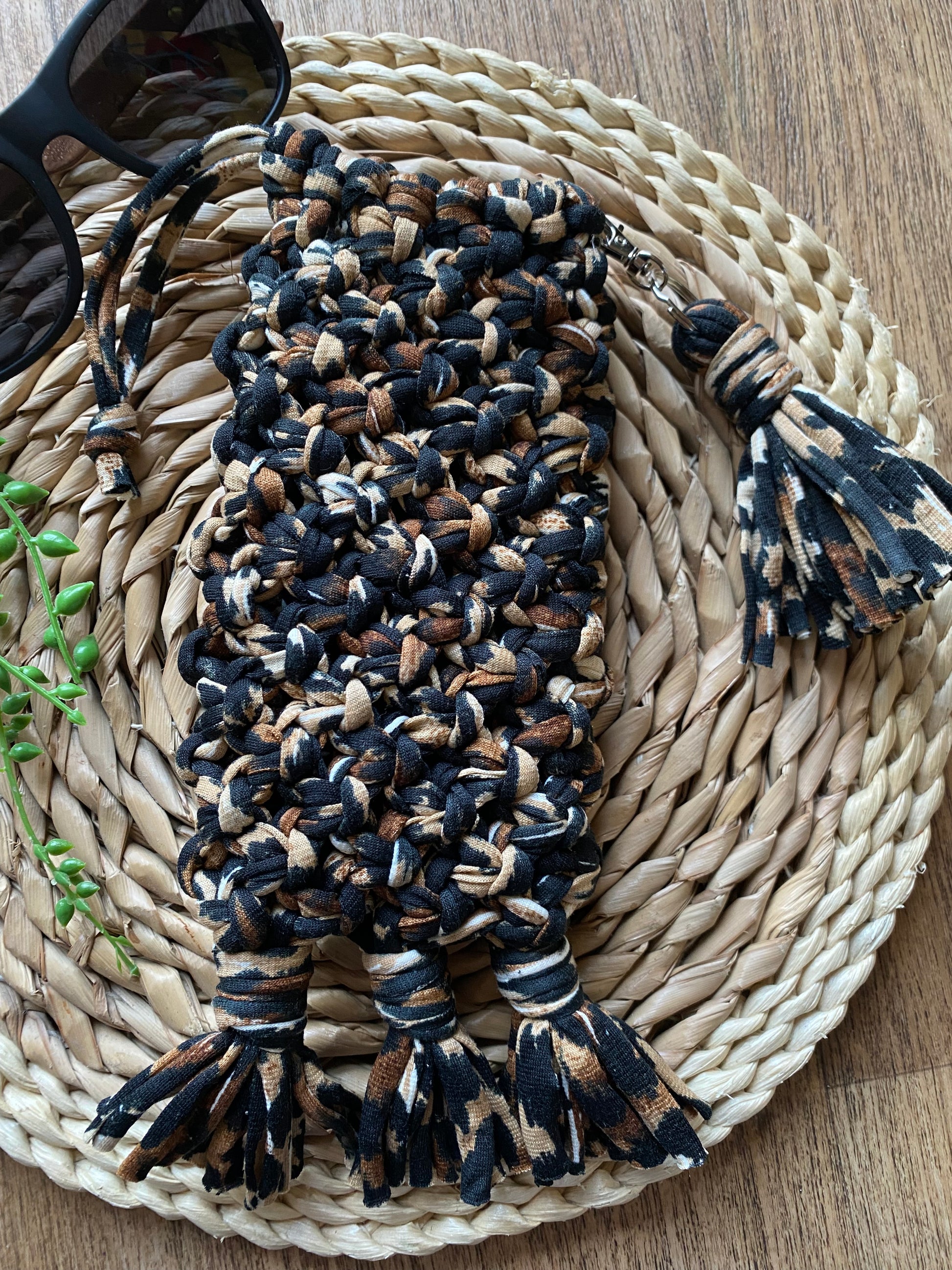 Handmade macrame soft glasses pouch made with animal print t shirt yarn. Displayed with on a round wicker mat, next to a dark pair of sunglasses.