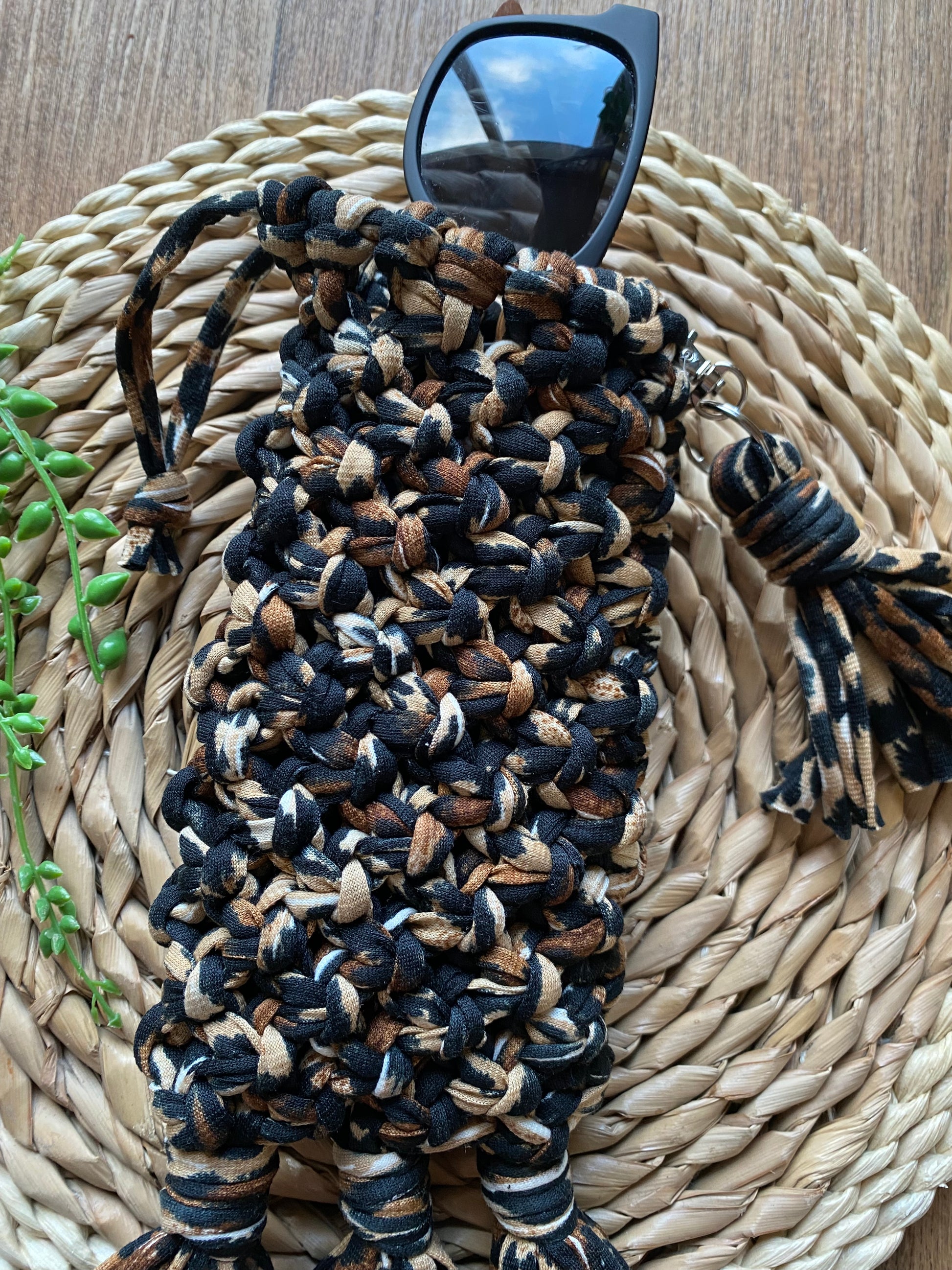 Handmade macrame soft glasses pouch made with animal print t shirt yarn. Displayed with a pair of dark sunglasses just showing out of the top of the pouch, on a round wicker surface.