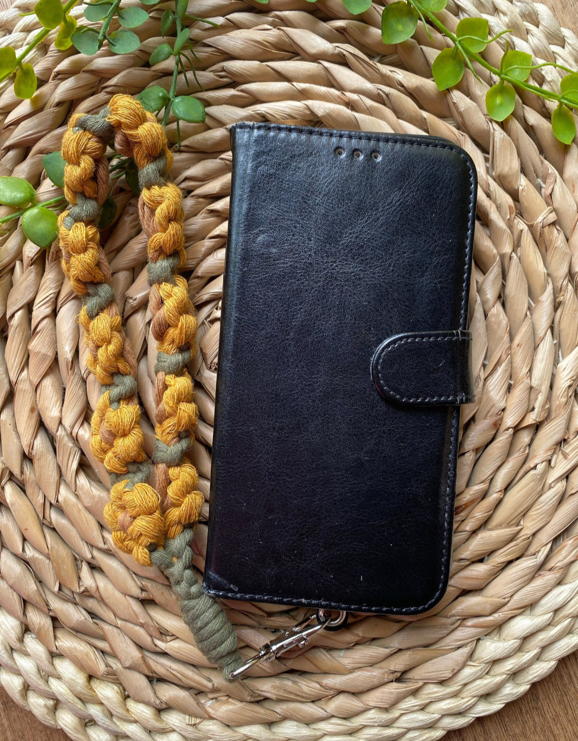A black leather phone case with a yellow and green sunflower-themed macramé phone charm wristlet attached to it, placed on a woven placemat with some green plant vines.
