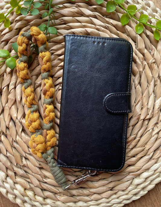 A black leather phone case with a yellow and green sunflower-themed macramé phone charm wristlet attached to it, placed on a woven placemat with some green plant vines.
