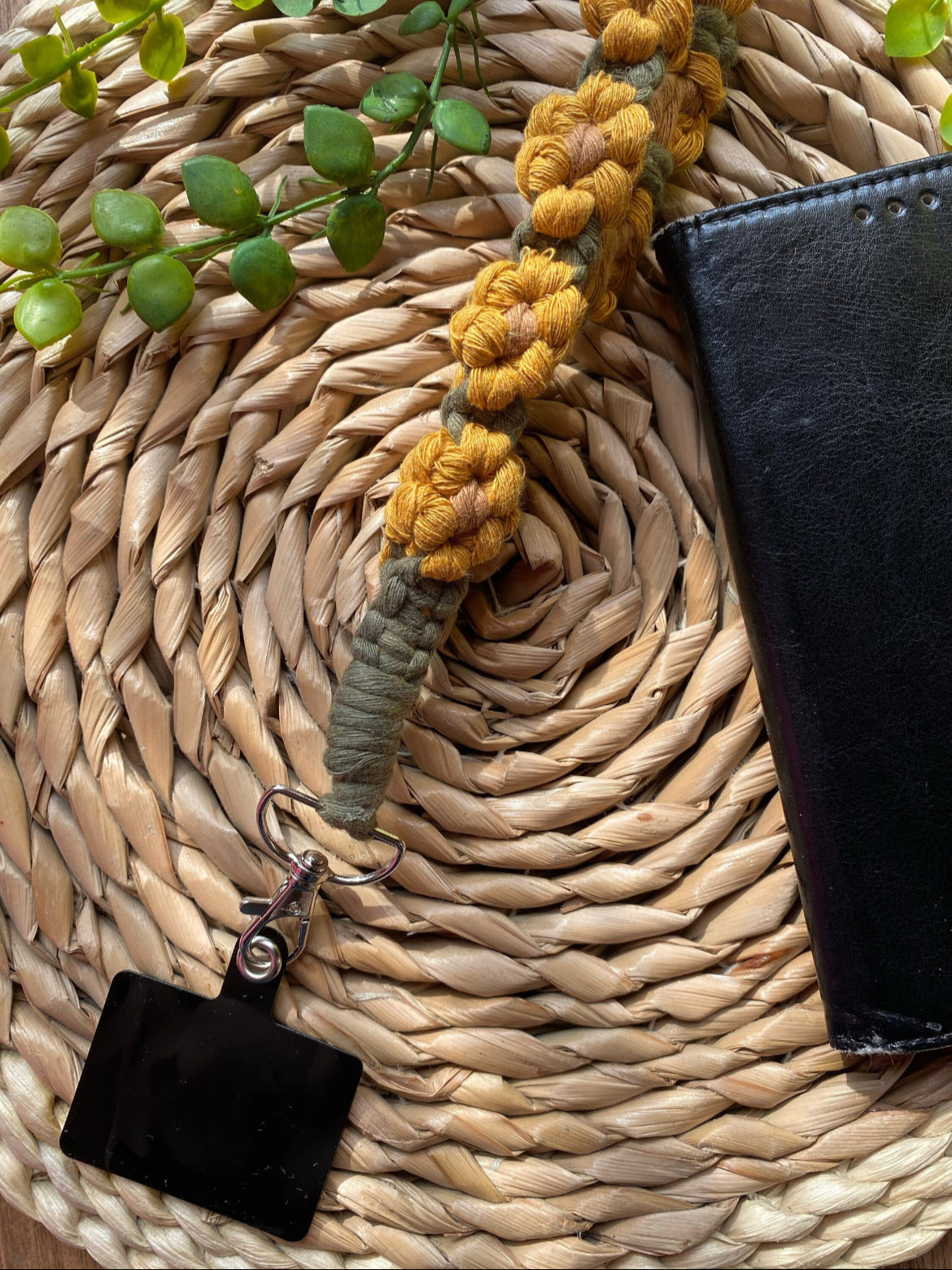 A yellow and green sunflower-themed macramé phone charm wristlet attached to a universal phone tether, with a metal clasp, on a woven mat with a black phone case and some green plant vines next to it.