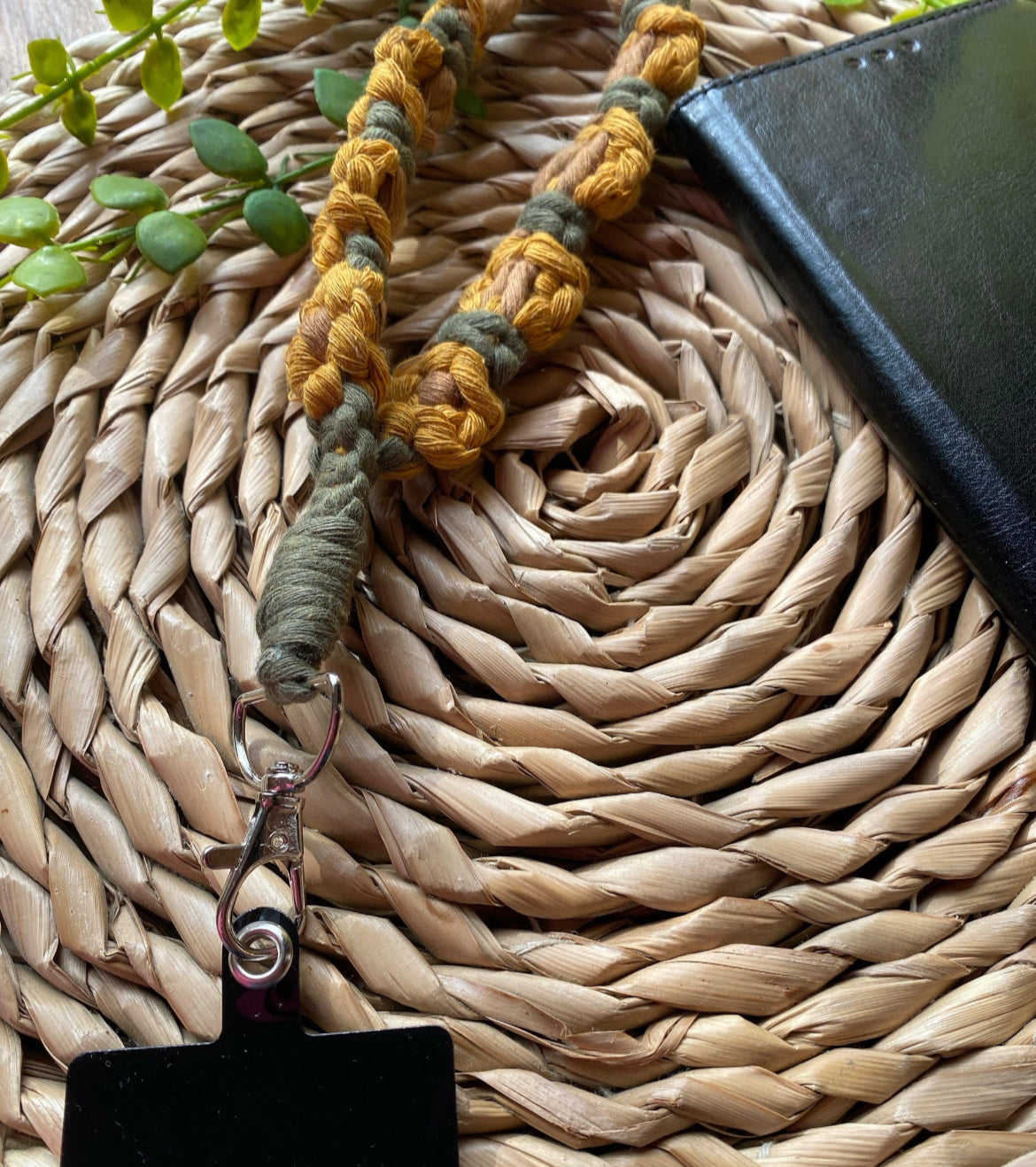 A close up image of a yellow and green sunflower-themed macramé phone charm wristlet attached to a universal phone tether with a metal clasp, on a woven mat.