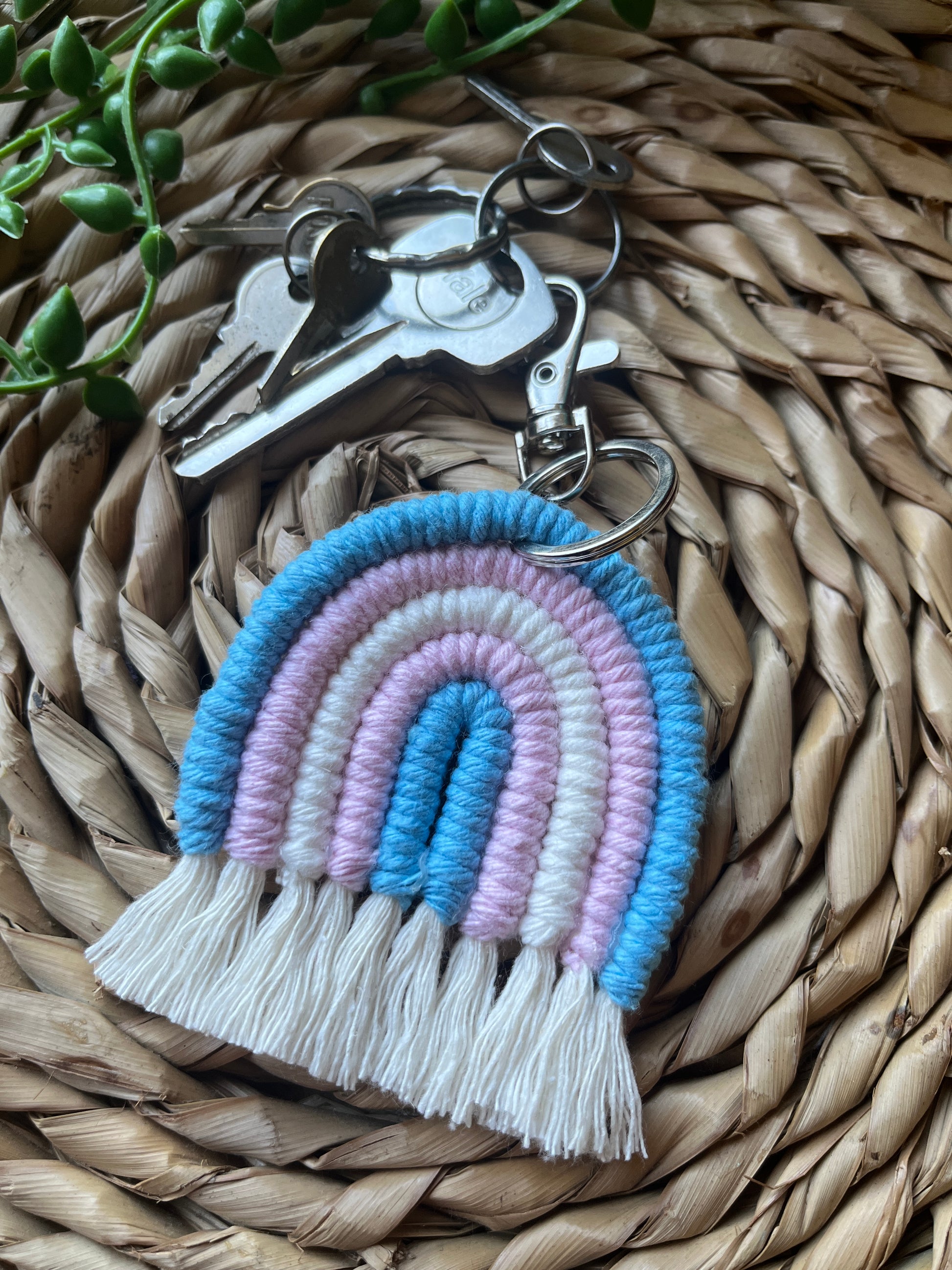 Handmade macrame rainbow keyring, made with blue, pink and white yarns to represent the colours of the trans pride flag. The keyring has a silver split ring and lobster clasp at the top, attached to a small set of silver keys.