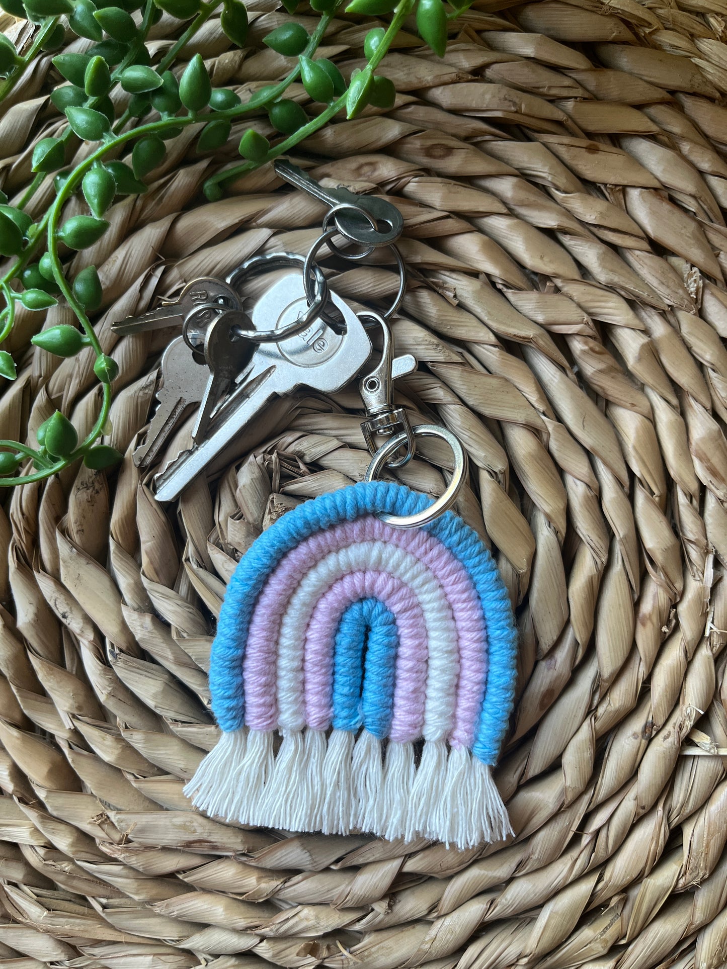 Handmade macrame rainbow keyring, made with blue, pink and white yarns to represent the colours of the trans pride flag. The keyring has a silver split ring and lobster clasp at the top, attached to a small set of silver keys.