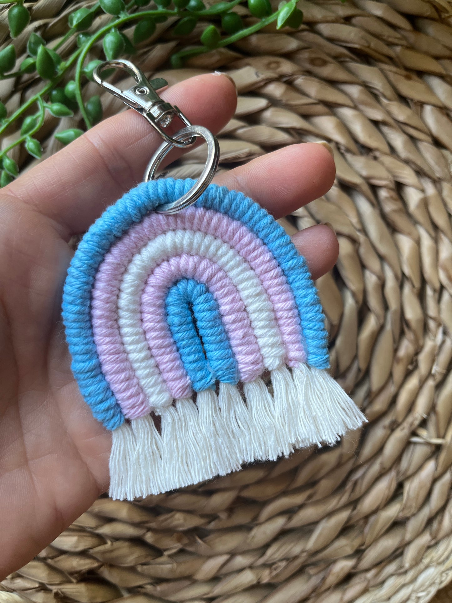 A hand holding a handmade macrame rainbow keyring, made with blue, pink and white yarns to represent the colours of the trans pride flag. The keyring has a silver split ring and lobster clasp at the top.