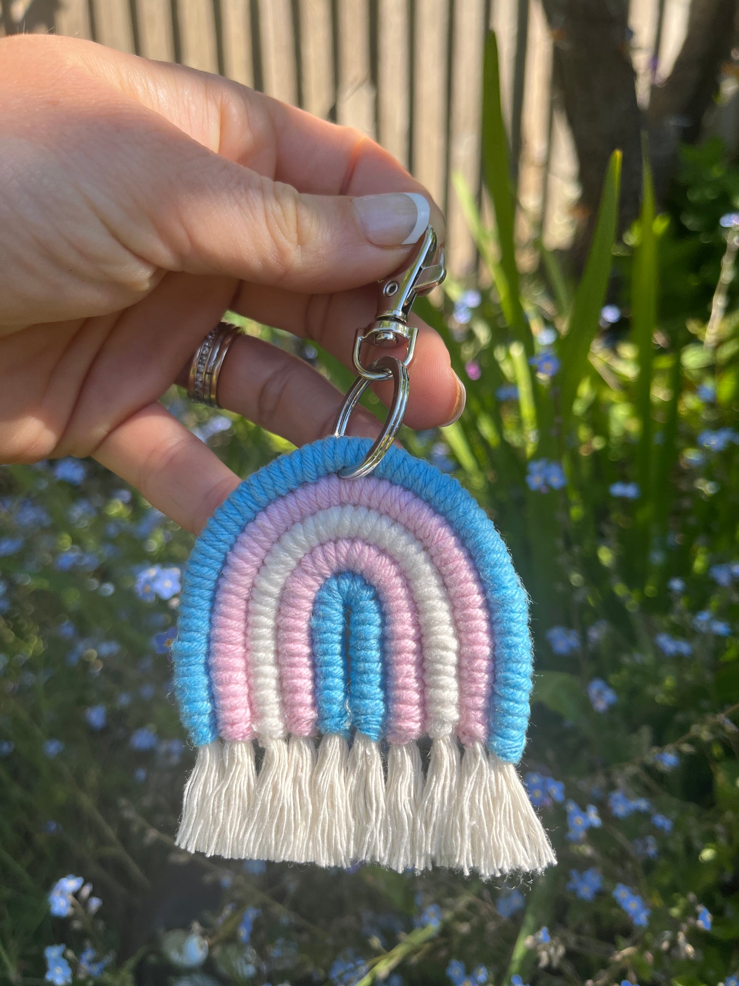 A hand holding a handmade macrame rainbow keyring, made with blue, pink and white yarns to represent the colours of the trans pride flag, outside in front of some forget me not flowers. The keyring has a silver split ring and lobster clasp at the top.