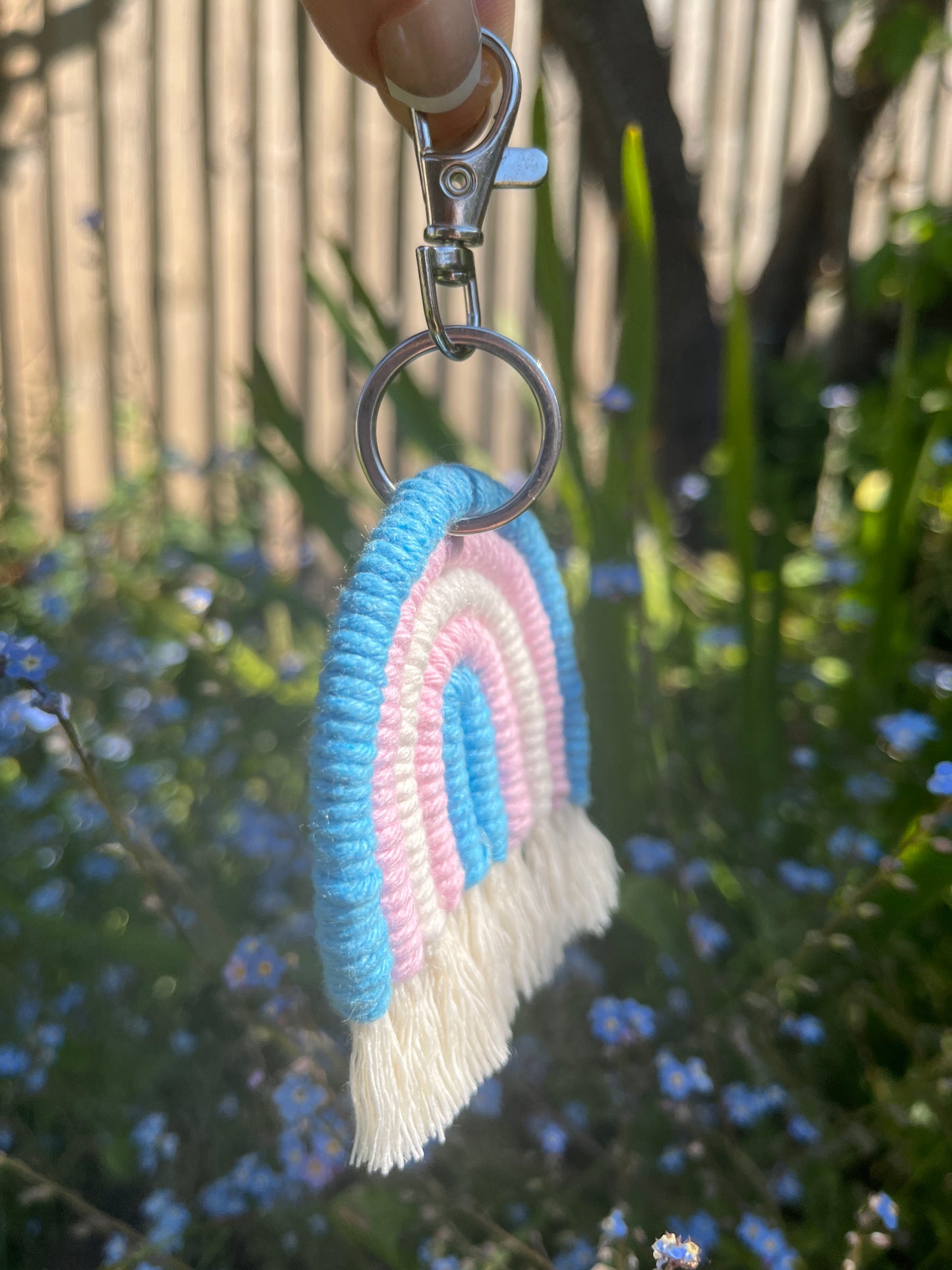 Handmade macrame rainbow keyring, made with blue, pink and white yarns to represent the colours of the trans pride flag. The keyring has a silver split ring and lobster clasp at the top.