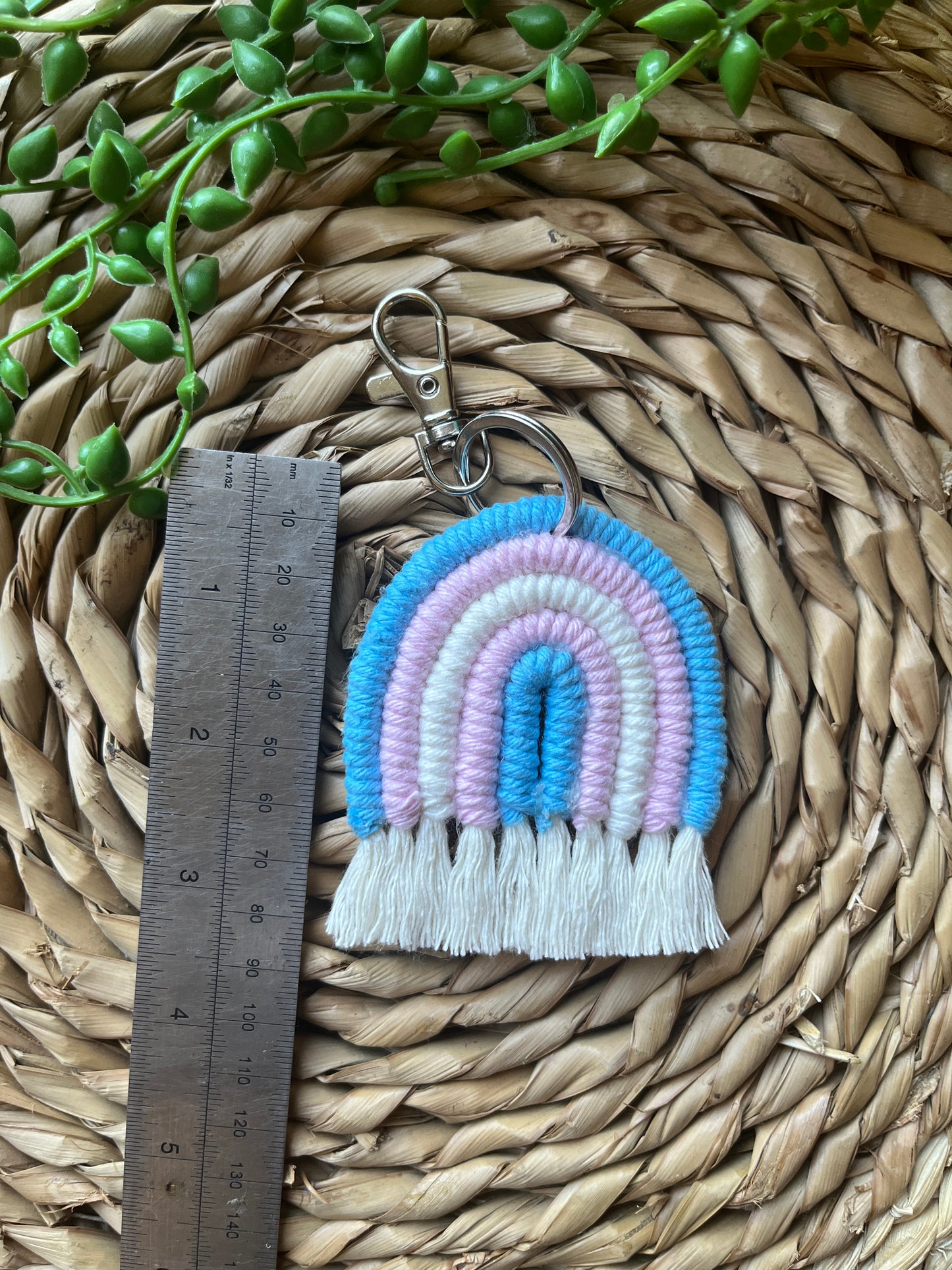 Handmade macrame rainbow keyring, made with blue, pink and white yarns to represent the colours of the trans pride flag. The keyring has a silver split ring and lobster clasp at the top. There is a metal ruler laid next to the keyring.