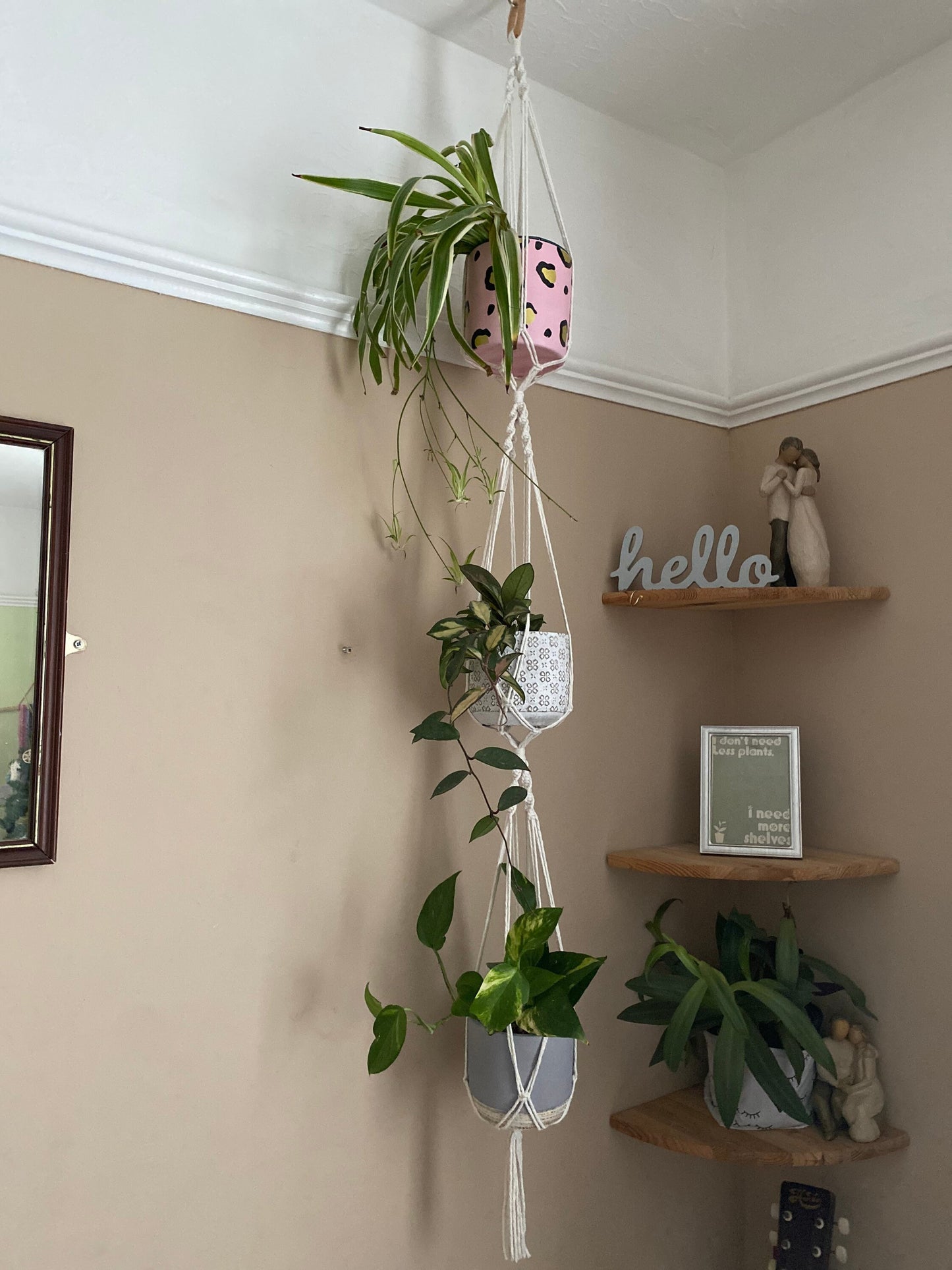 Three large green plants in ceramic plant pots, hang vertically in a handmade macrame triple plant hanger, made with natural coloured yarn, suspended from the ceiling with a wooden ring.