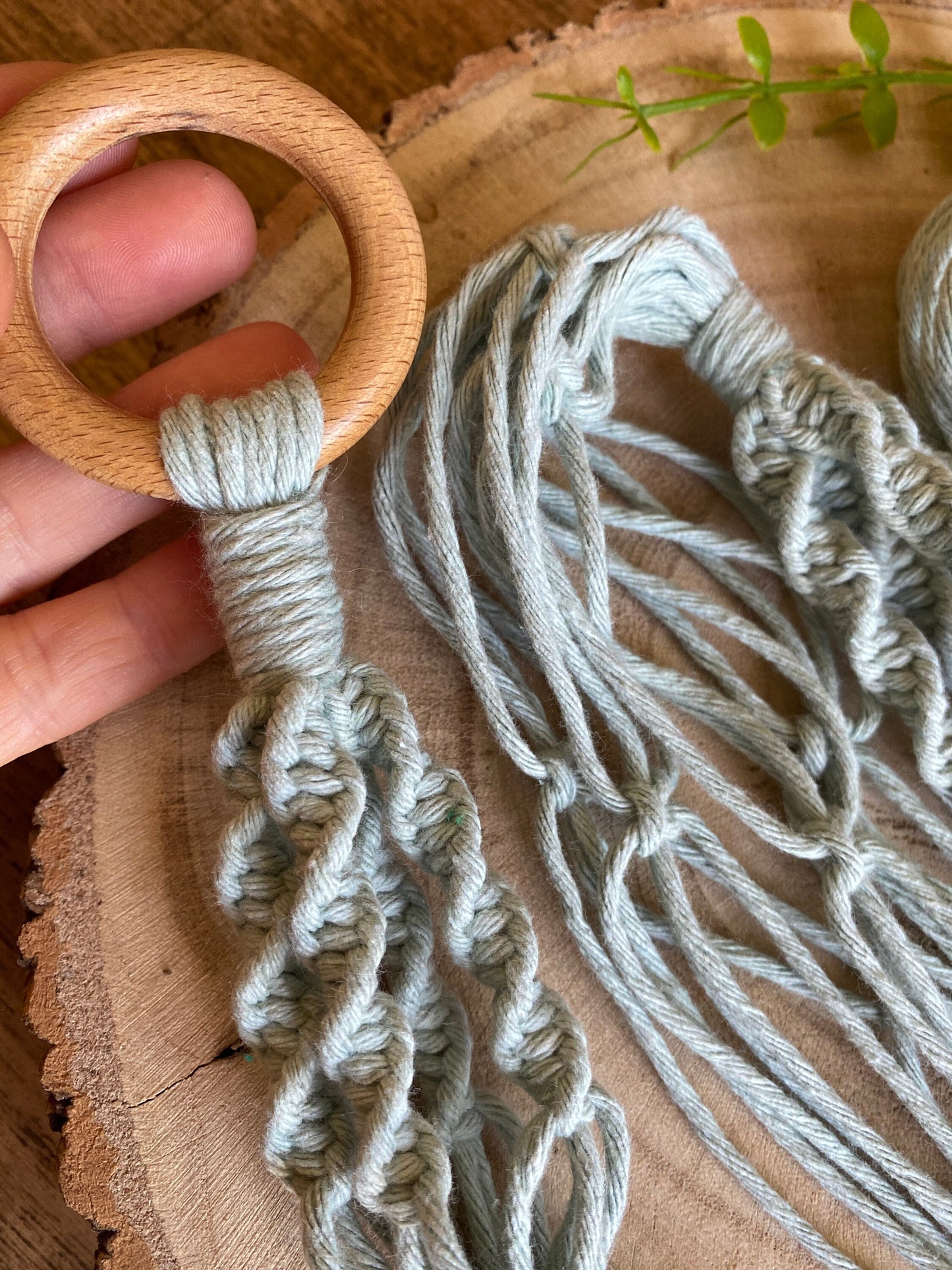 A hand holding the wooden ring at the top of a handmade macrame plant hanger, made with a twisted knot pattern in a pastel green yarn.