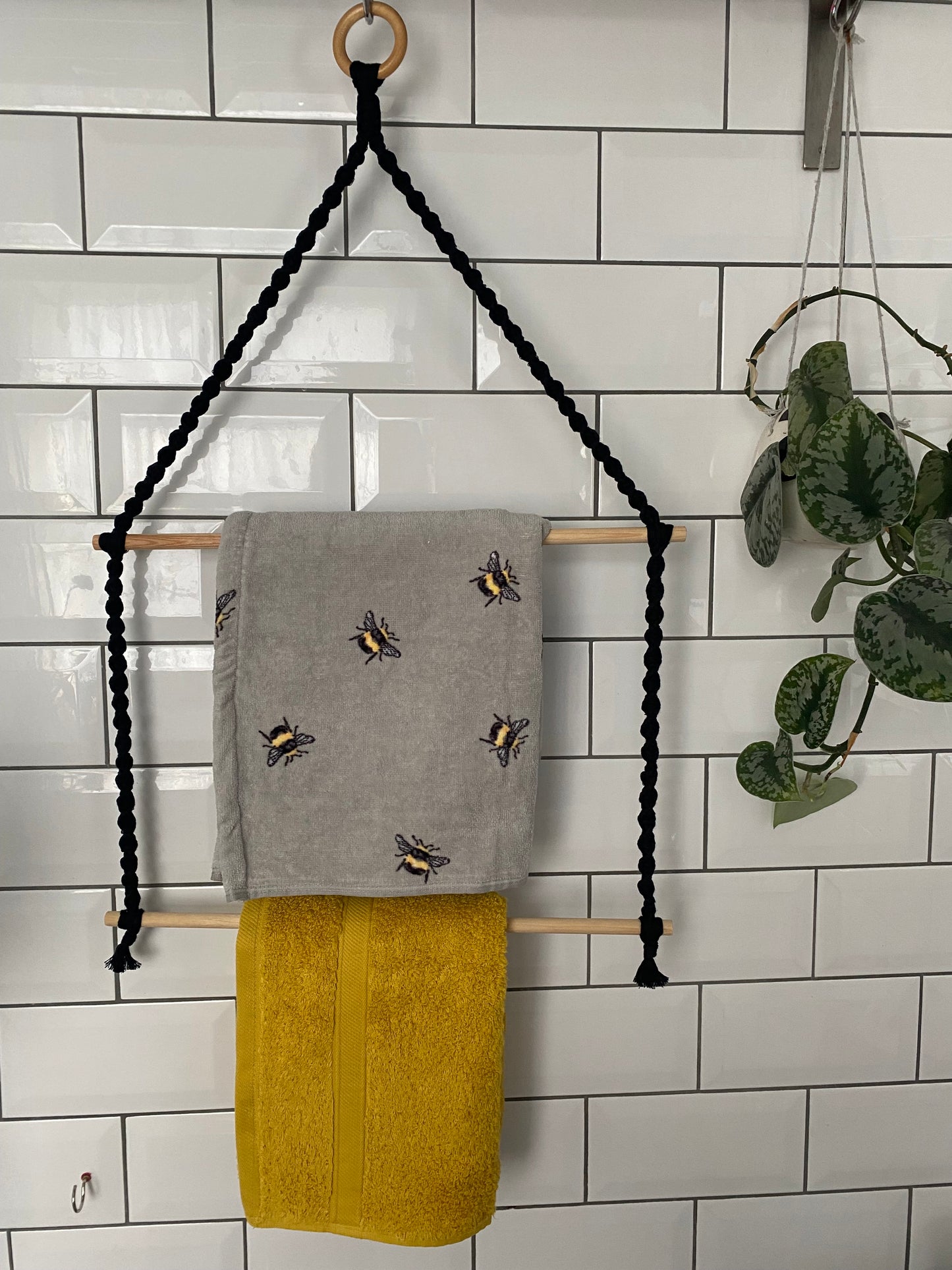 A handmade towel rack made with black yarn, a wooden ring and two wooden dowels, holding a grey towel with bee pattern and a yellow towel, hangs against a white tiled wall.
