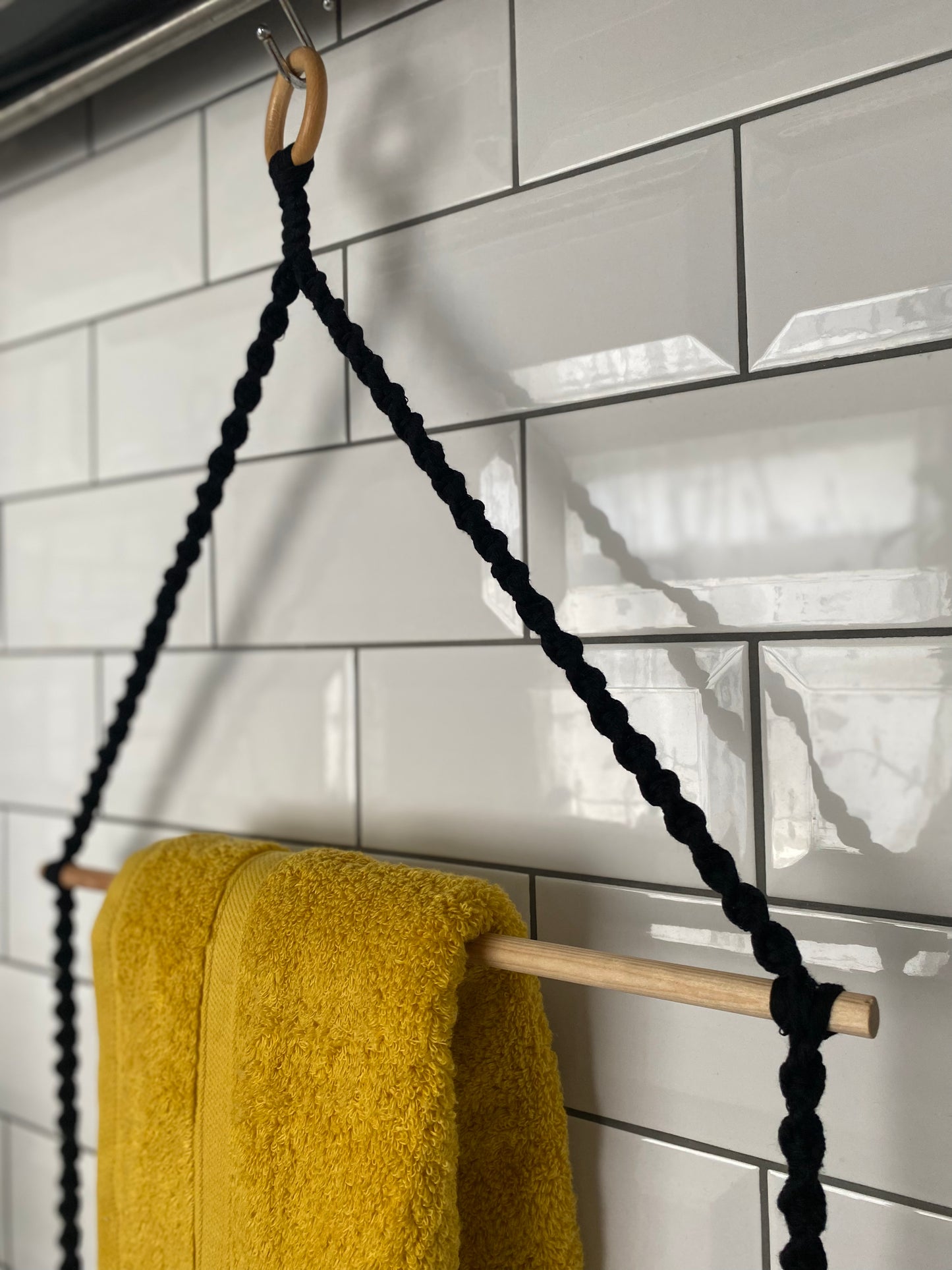 Close up of a macrame towel rack, made with black yarn, a wooden ring and wooden dowels, holding a yellow towel, hanging against a white tiled wall.