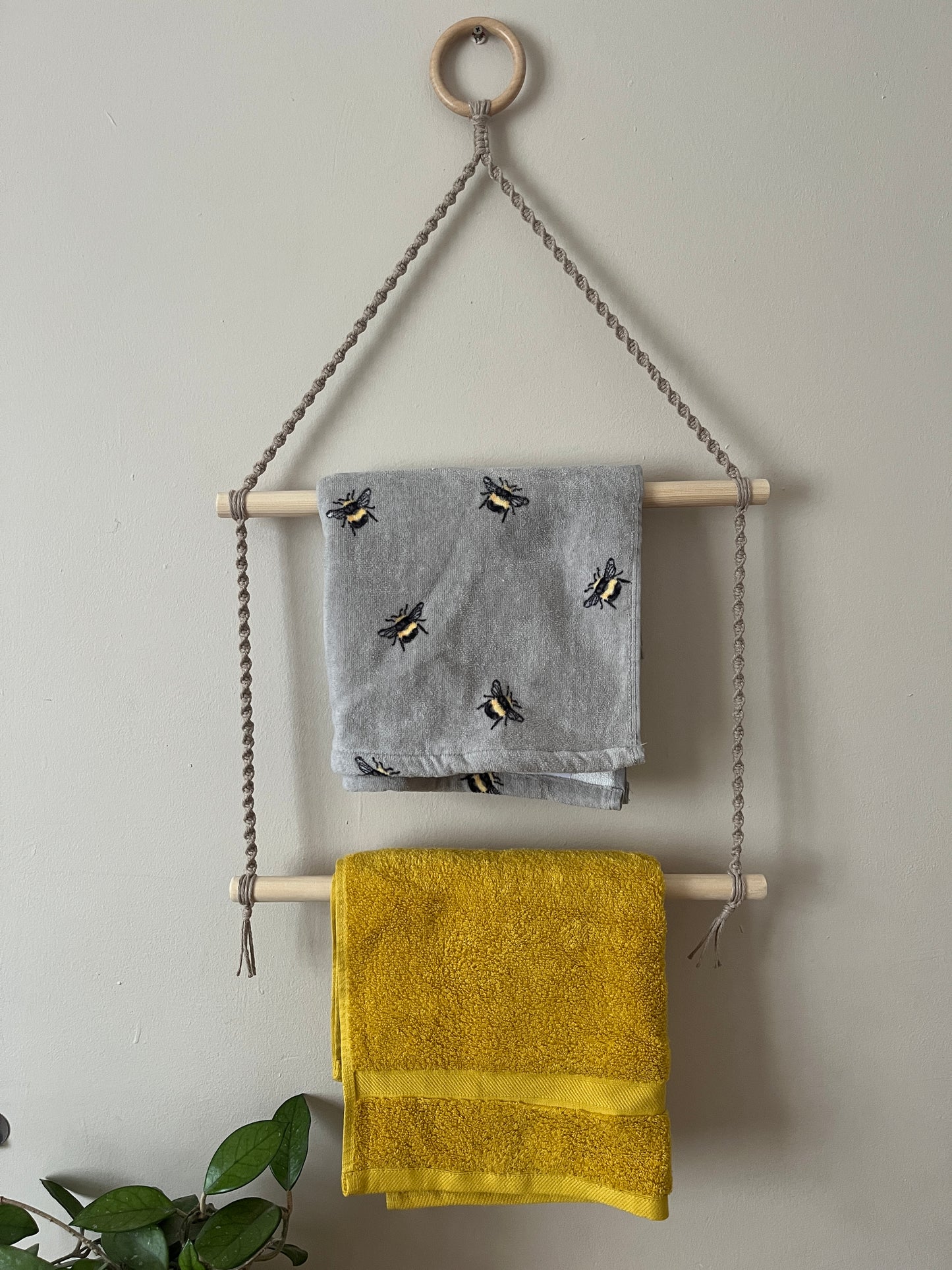 Two towels hanging on a handmade towel rack, made with taupe yarn, wooden dowels and a wooden ring against a plain wall.