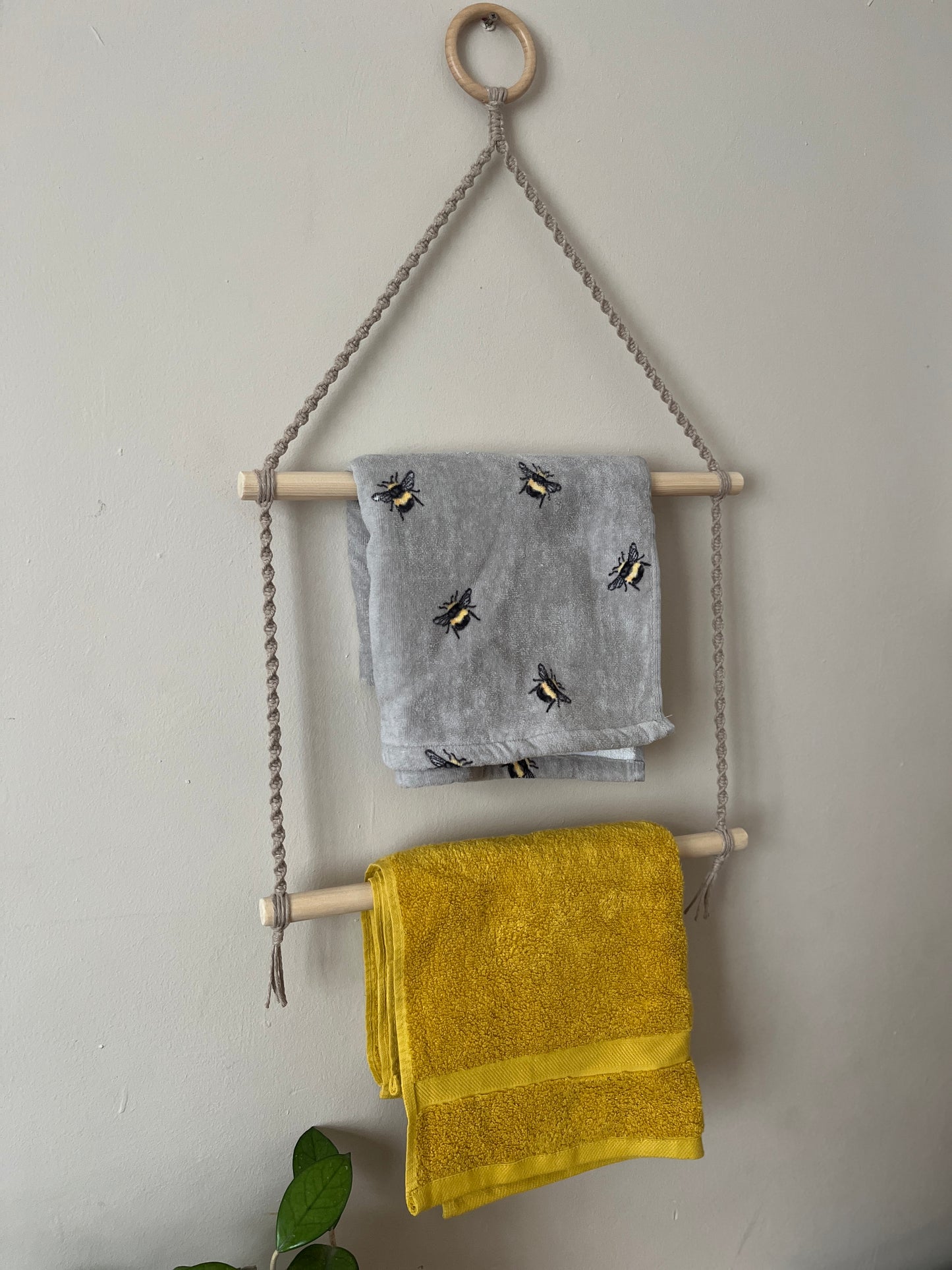 Towel rack made with taupe yarn, a wooden ring and wooden dowels, with grey and yellow towels hanging on it, against a plain wall