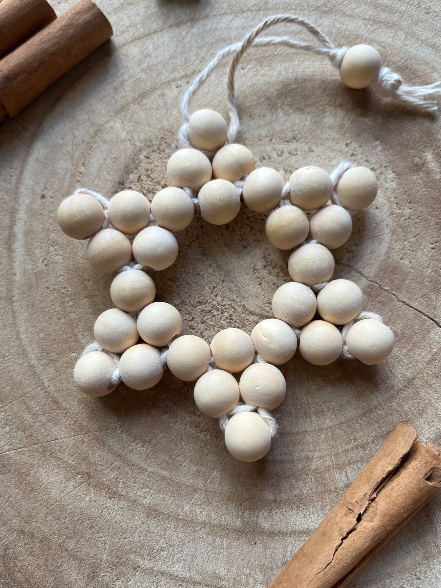 Close up of a star-shaped hanging decorative item made of small wooden beads and cotton yarn, on a wooden surface with cinnamon sticks and gold beads for decoration.